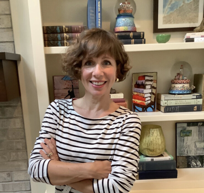 A woman with short curly hair smiling and standing with her arms crossed in front of a bookshelf filled with books and decorative items.
