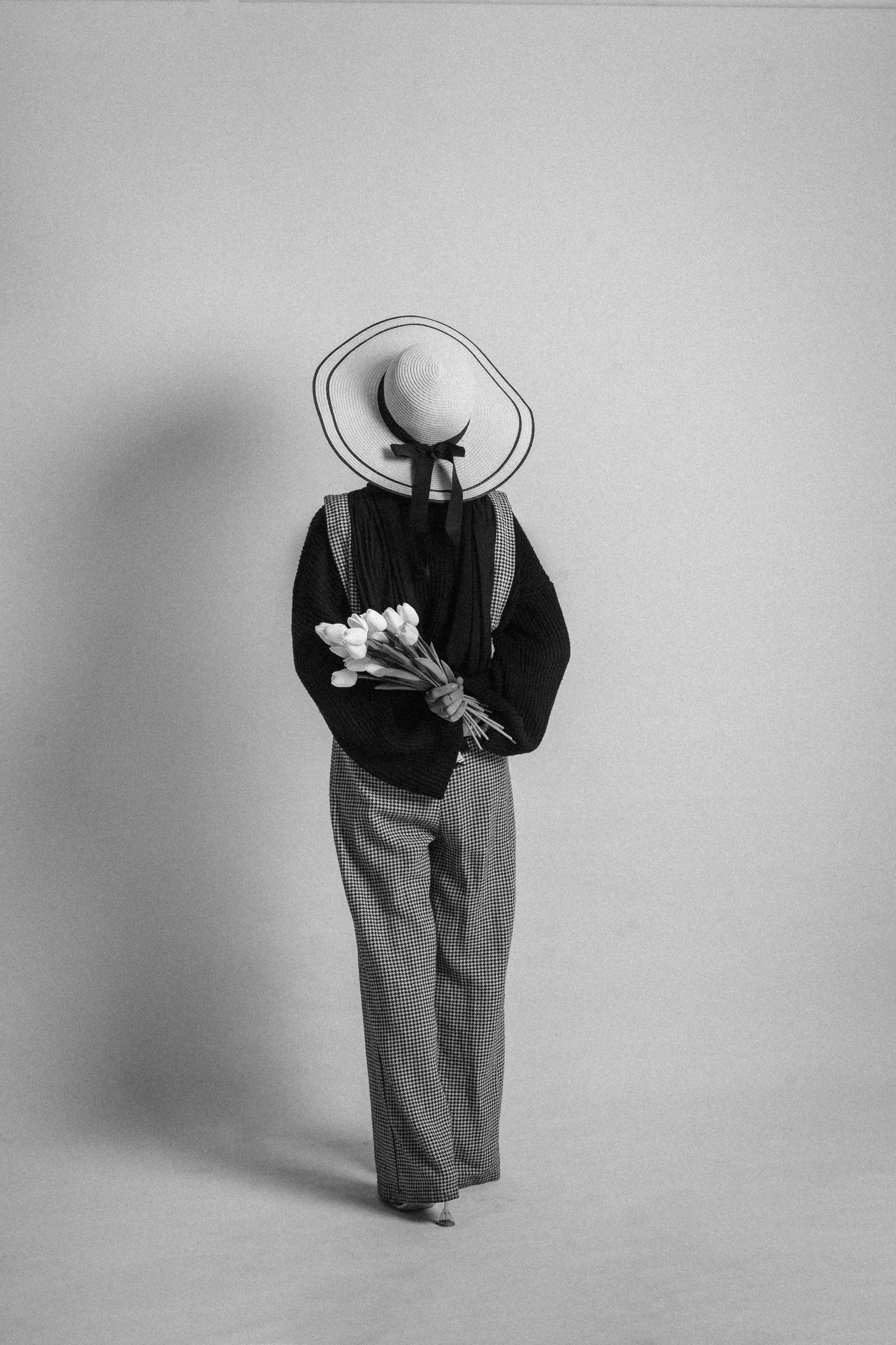 A person standing against a plain wall, wearing a large sunhat with a ribbon, a dark sweater, checkered pants, and holding a bouquet of white tulips.