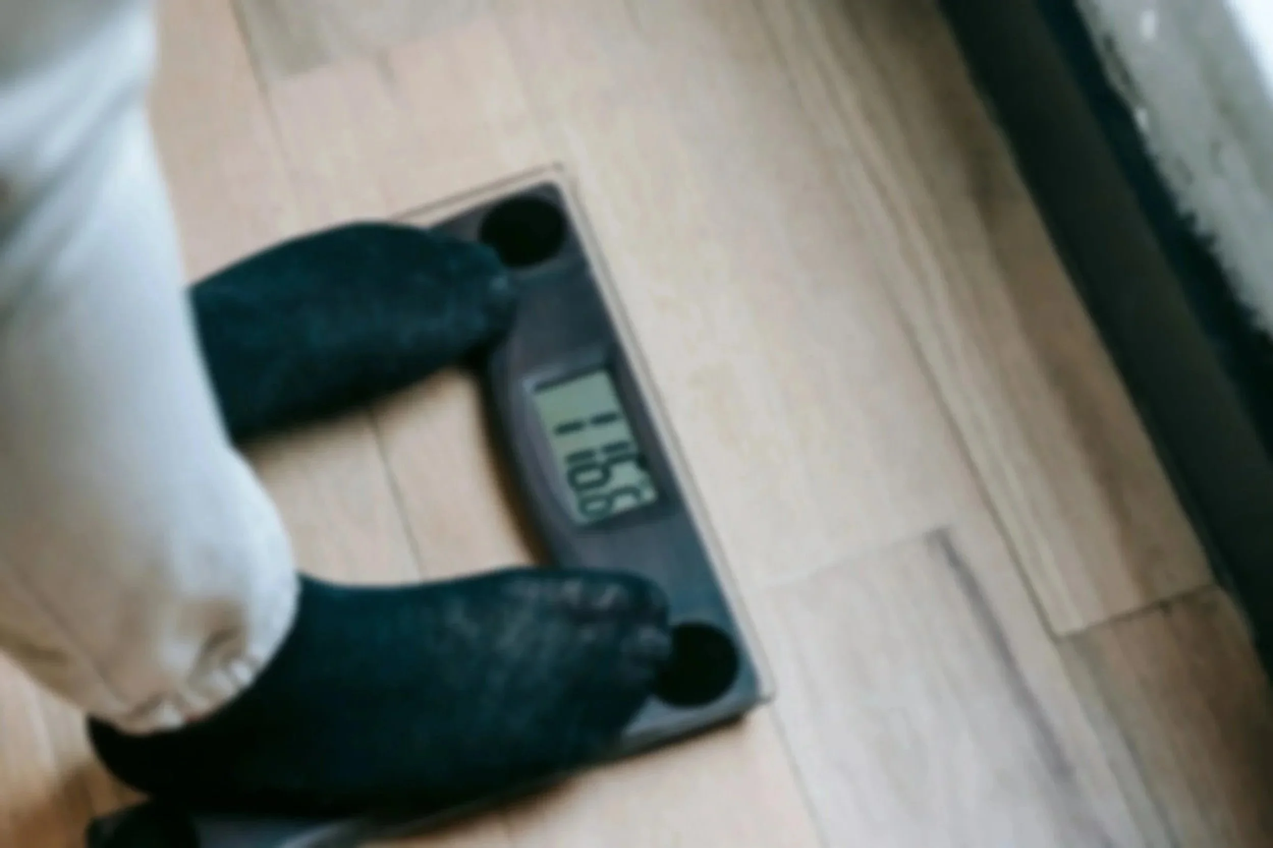 Person standing on a digital bathroom scale with black socks and light-colored pants, the scale shows a weight of 158.9 pounds, on a wooden floor.