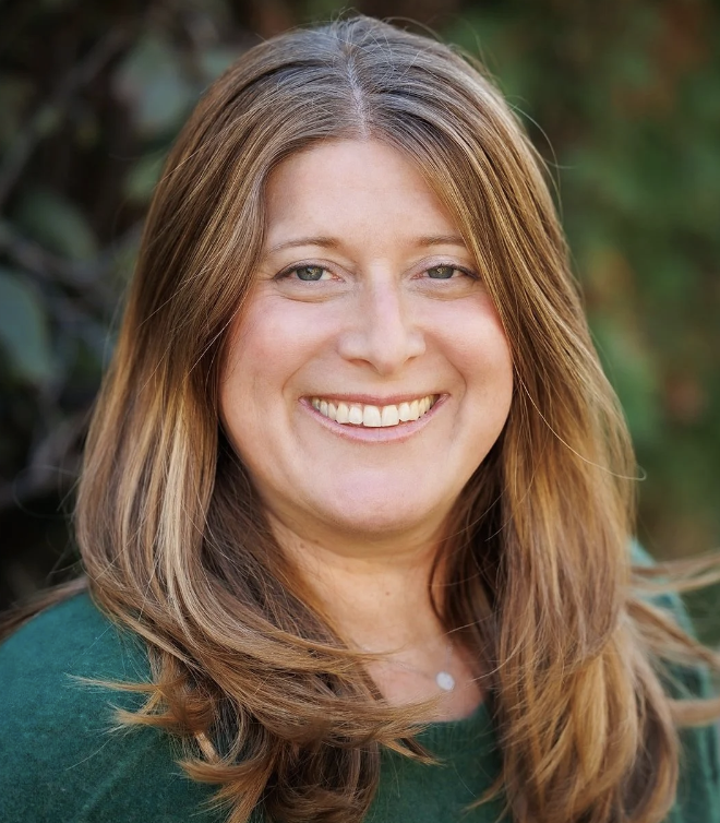 A woman with shoulder-length brown hair, smiling outdoors with greenery in the background.