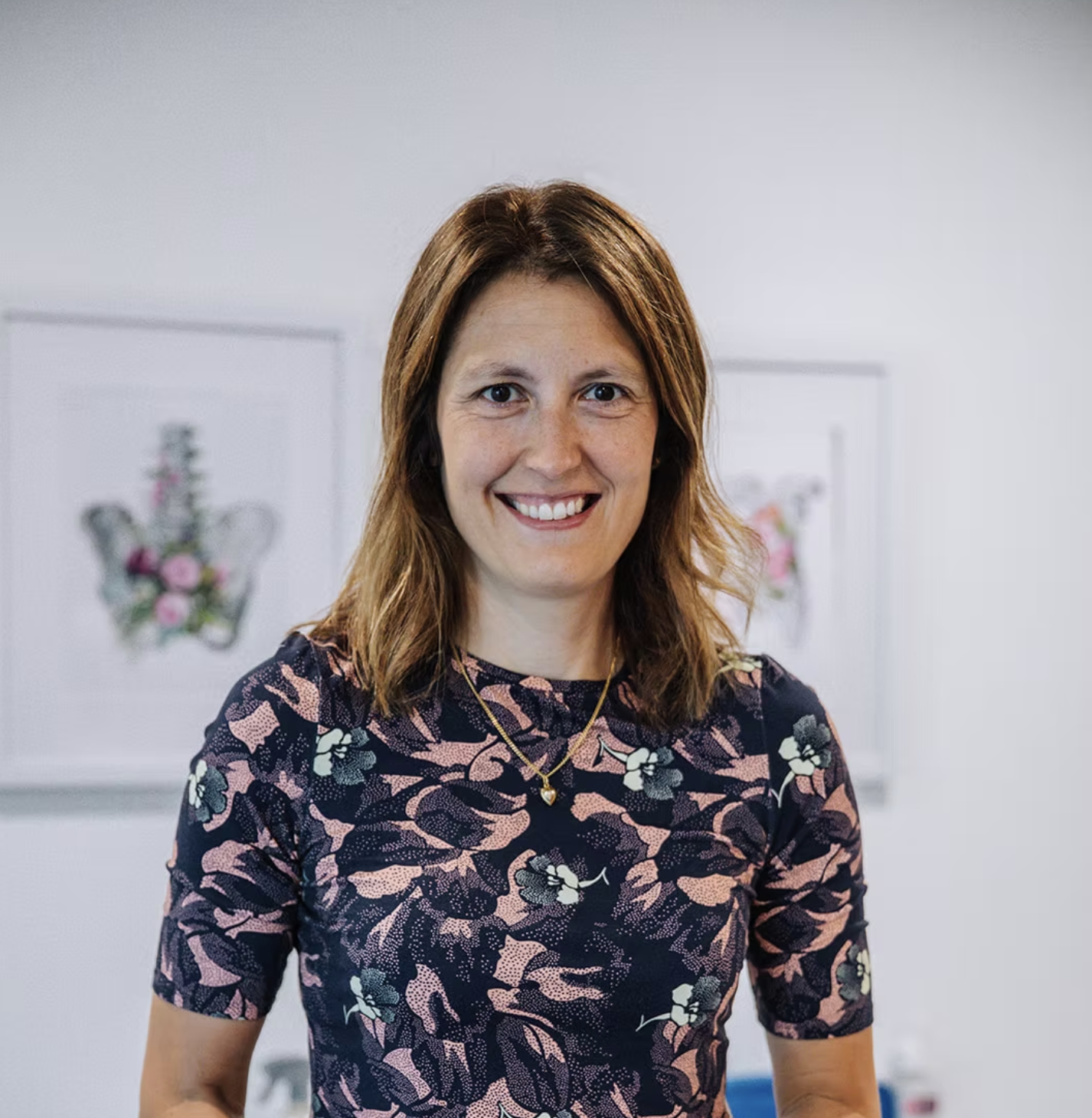 A woman with shoulder-length brown hair smiling, wearing a floral-patterned dress and a gold necklace, standing in front of framed floral art.