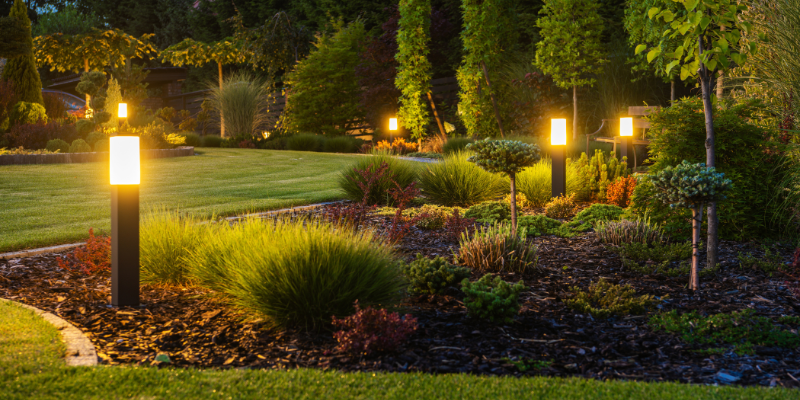 Nighttime garden with illuminated pathway lights, shrubs, trees, and manicured lawn.