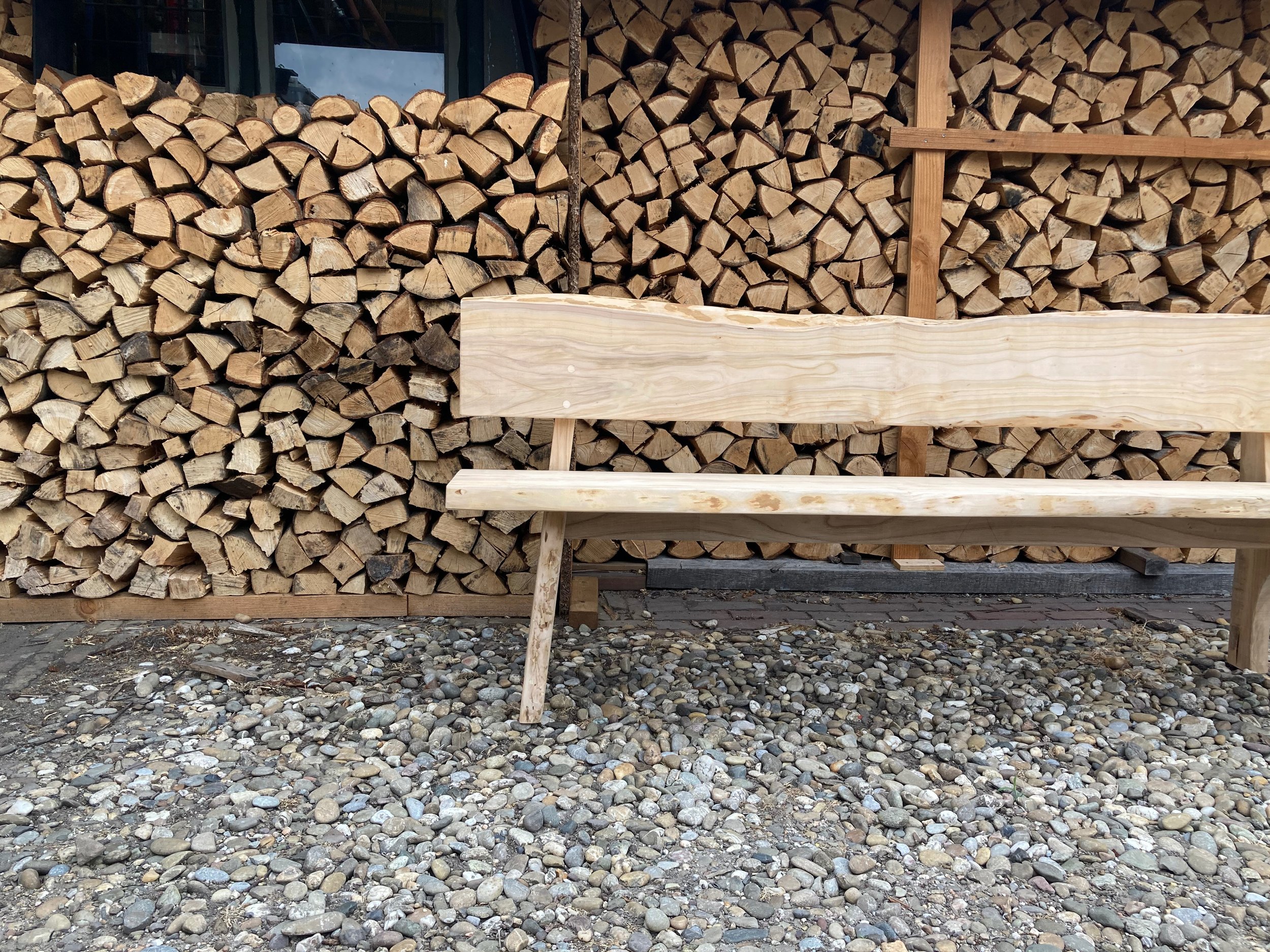 Handgemaakte boomstam bank van massief hout, ontworpen door Tije Interieur. Robuuste houten tuinbank met natuurlijke randafwerking, geplaatst voor een houtopslag met gekloofd brandhout in Zutphen (Gelderland).
