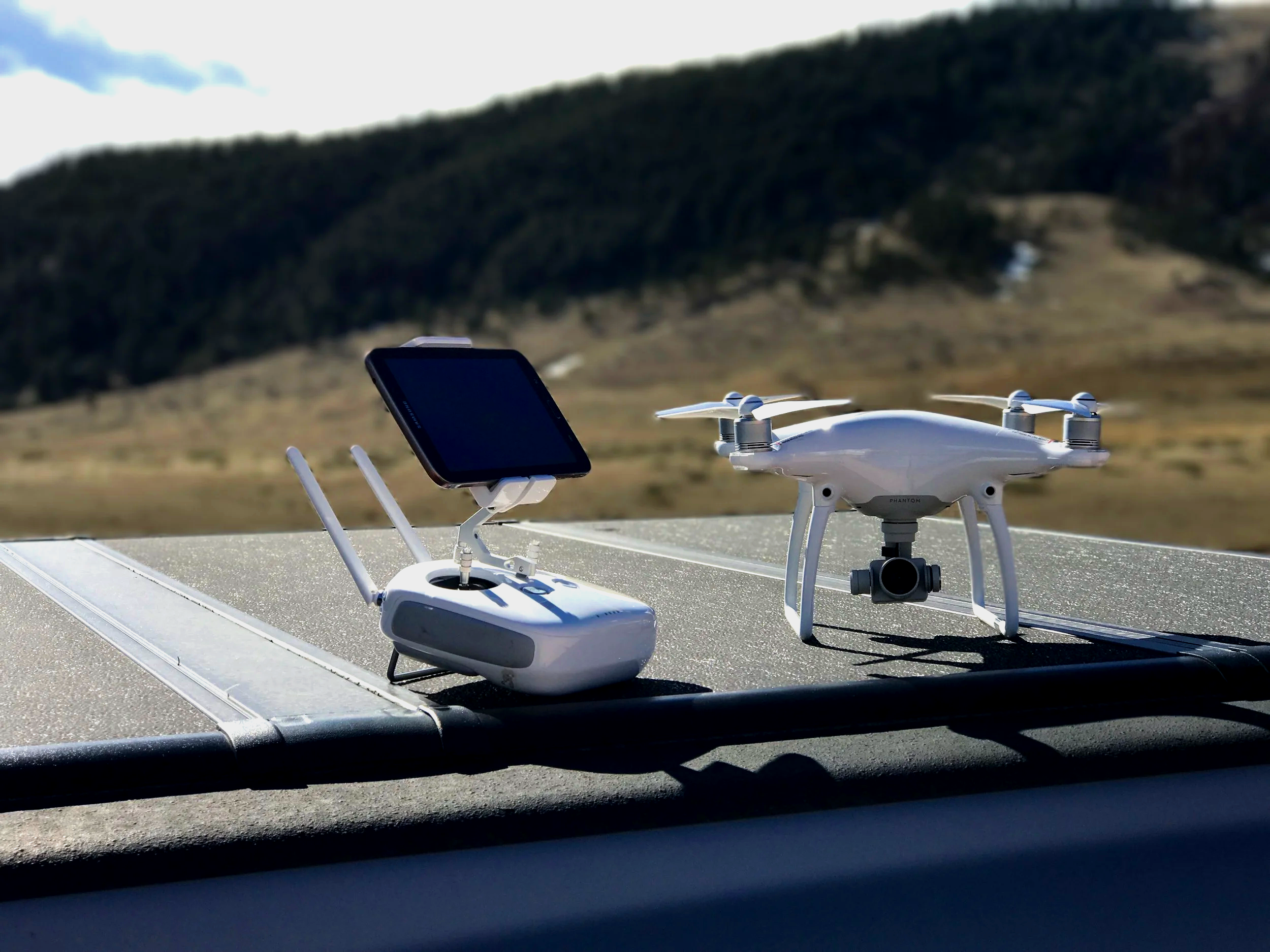 A drone with a mounted camera and a remote controller with a screen, set on a surface outdoors with grassy hills in the background.