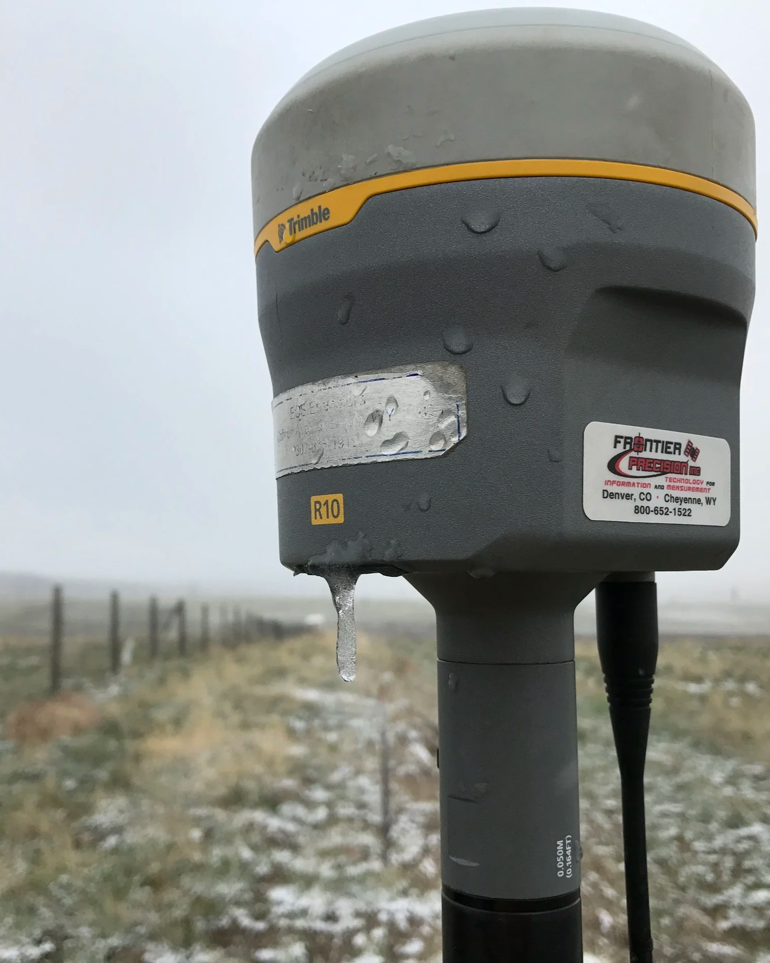 Close-up of a surveying instrument with water droplets and ice, set in a foggy outdoor landscape with a fence and snow-covered ground in the background.