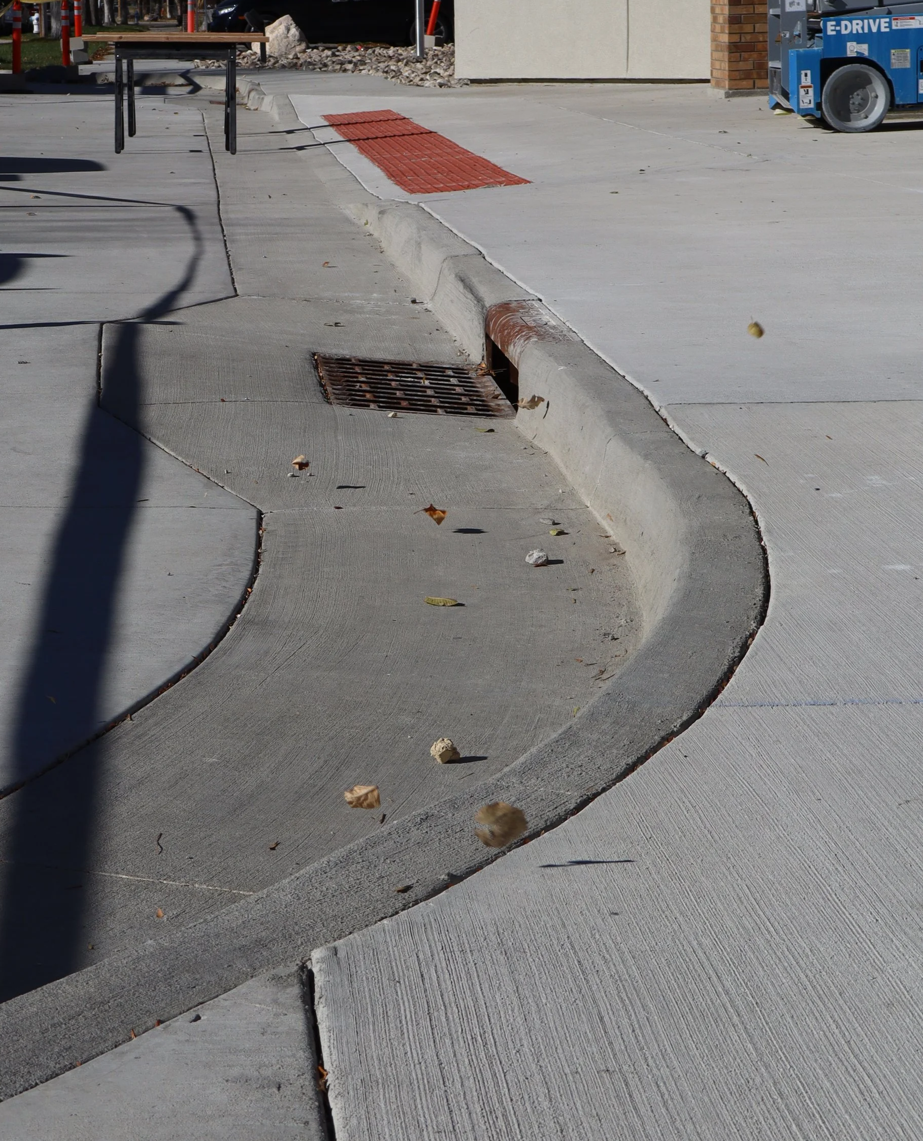 Sidewalk with a curb and drain grate, leaves scattered on the ground, part of a blue lift in the background.