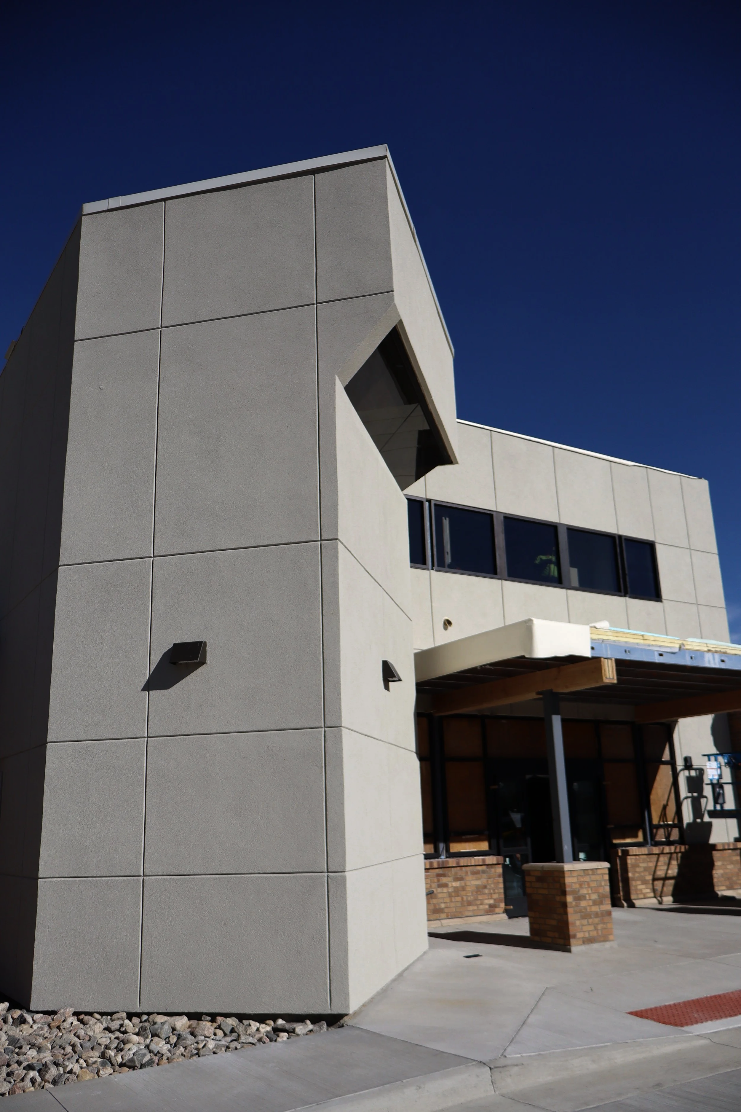 Modern building with beige concrete walls, black-framed windows, and a brick base, under a clear blue sky.
