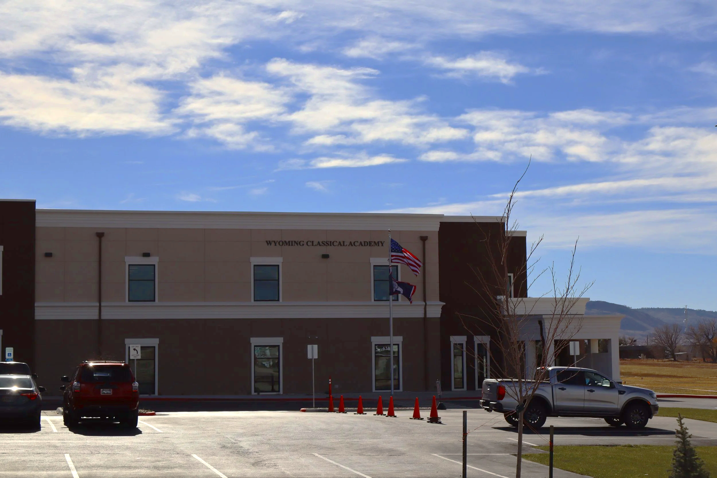 Front view of Wyoming Classical Academy building with parking lot, American flags, some trees, and mountains in the background under a partly cloudy sky.