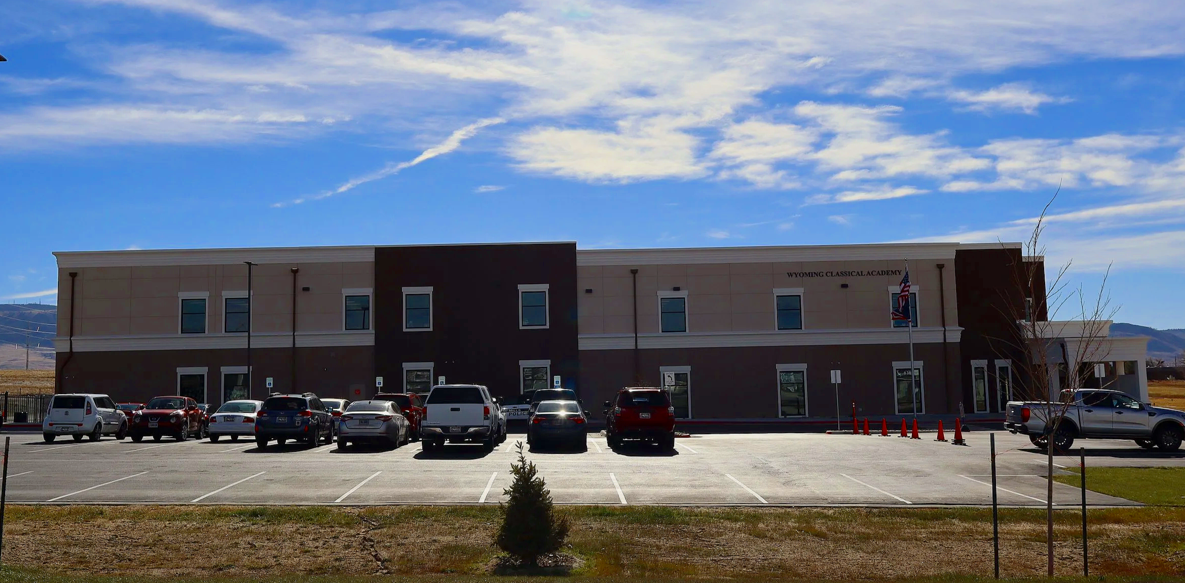 Front view of a modern two-story building with a sign reading 'Wyoming Classical Academy', parked cars in the foreground, a tree, and a blue sky with clouds in the background.