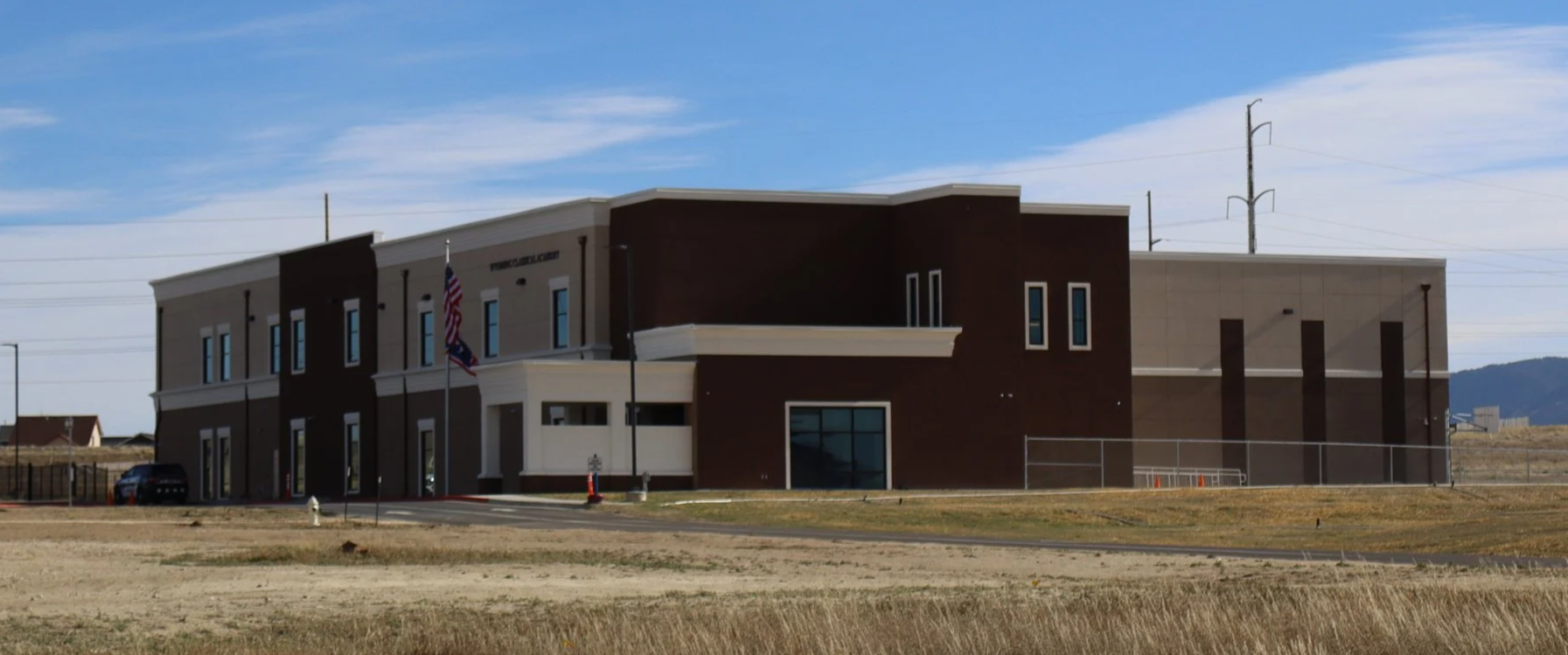 Modern two-story building with brown and beige exterior, large windows, and American flags, situated in an open field with a clear blue sky.