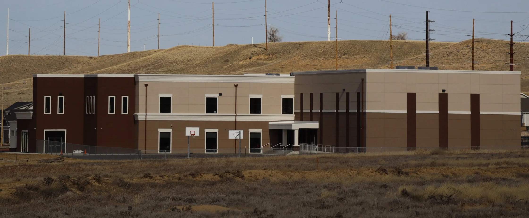 Modern school building with multiple windows, basketball hoops outside, and a fence, set against hilly terrain with utility poles in the background.
