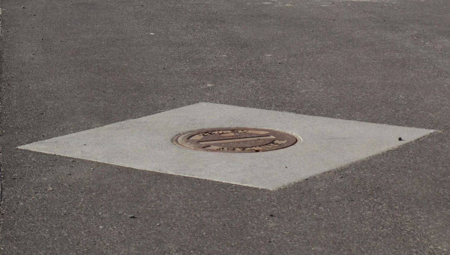 A manhole cover embedded in asphalt pavement, surrounded by a painted white square boundary.