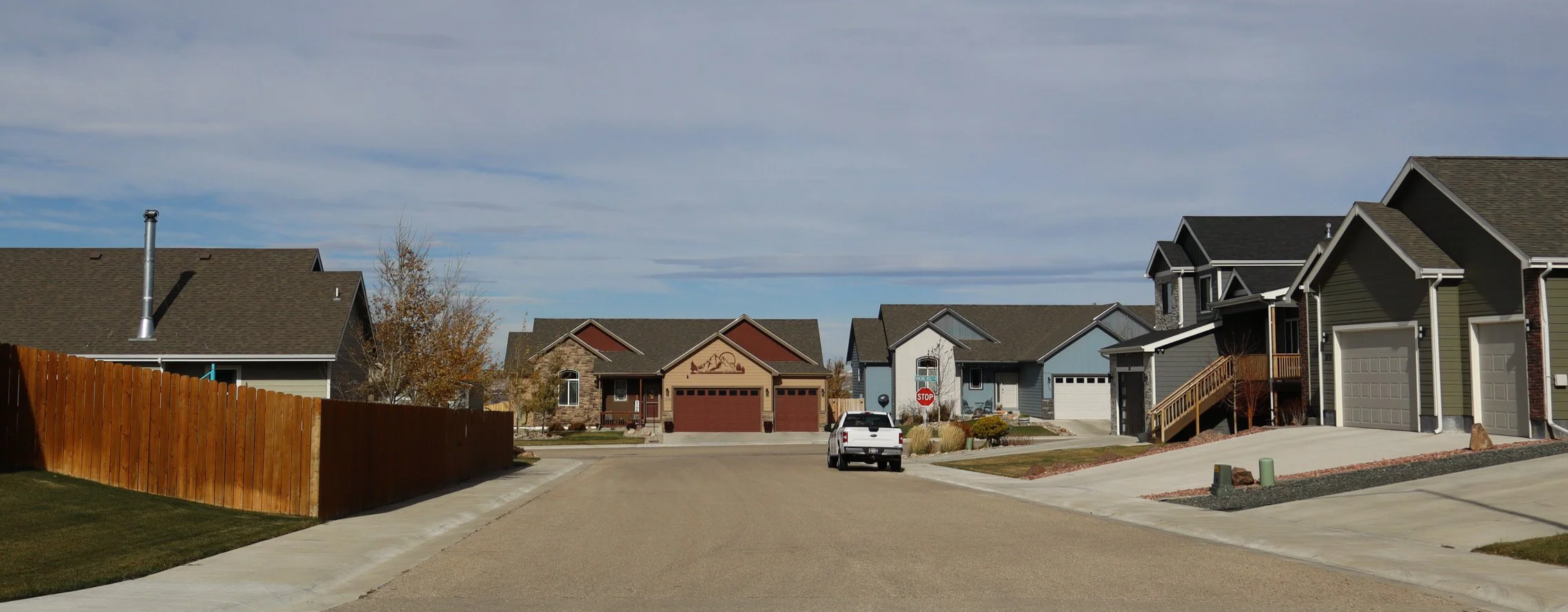A residential neighborhood with multiple two-story houses, a street, parked cars, a stop sign, a wooden fence, and a clear sky.