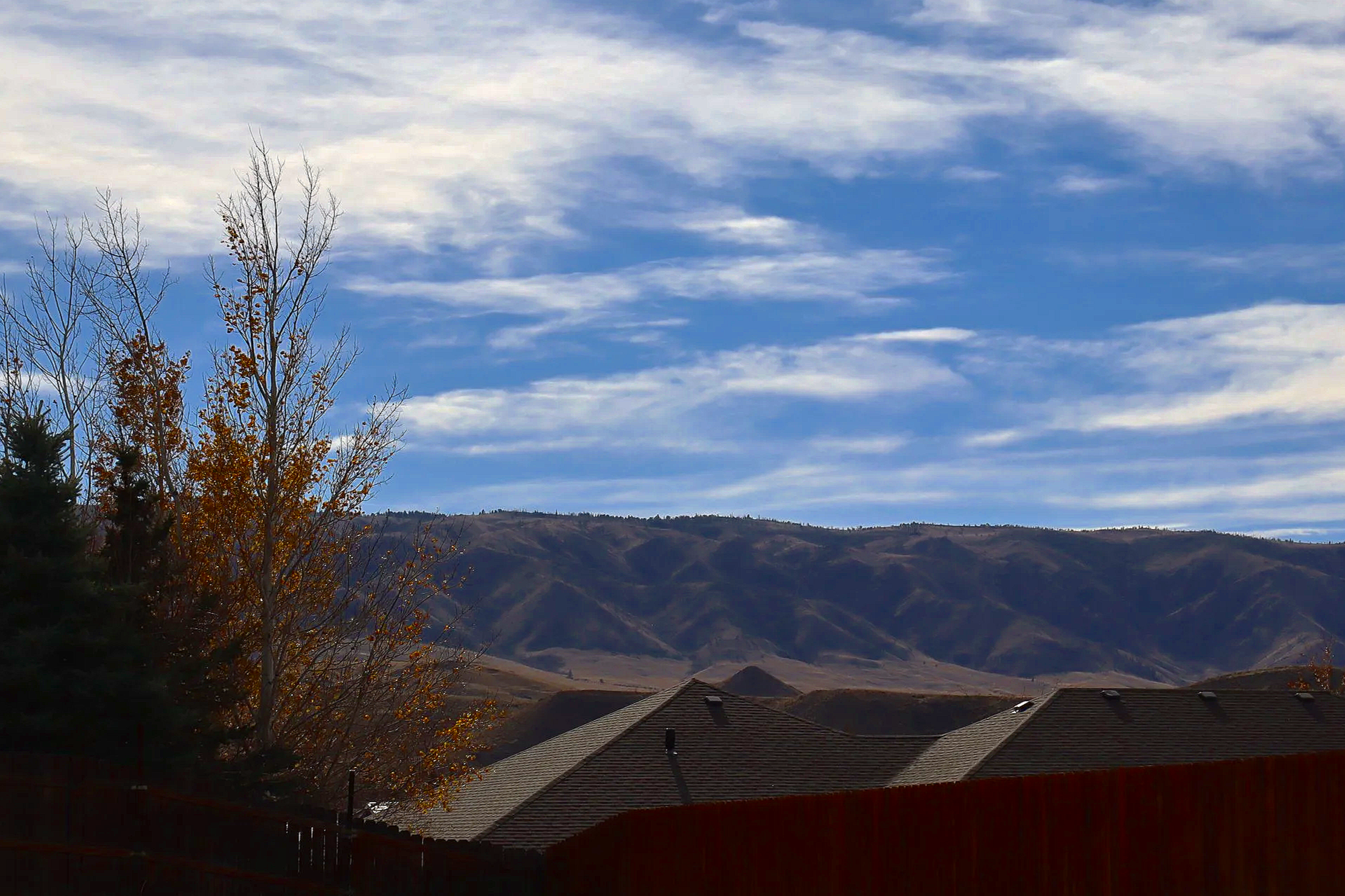 Houses with sloped roofs in front of mountains and a sky with scattered clouds, trees with some leaves changed to fall colors, and a wooden fence in the foreground.