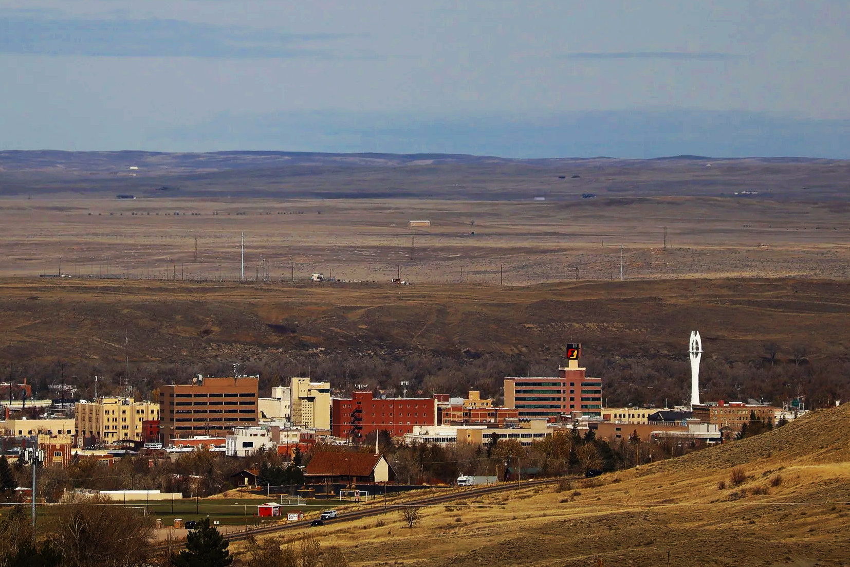 A cityscape with various buildings including a tower and a sculpture, surrounded by open countryside and hills in the background.