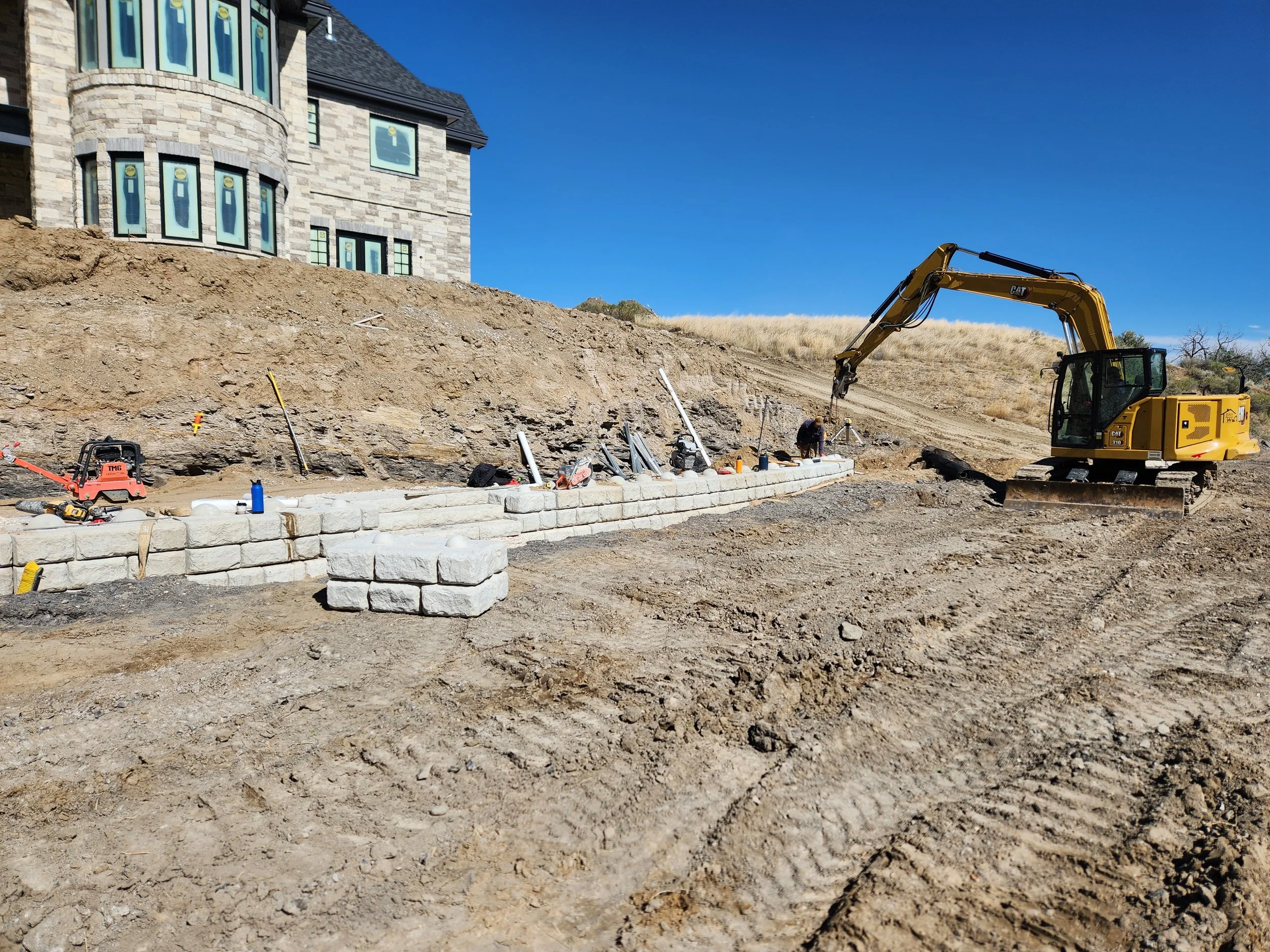 Construction site with a yellow excavator building a stone wall in front of a house on a hill, under a clear blue sky.