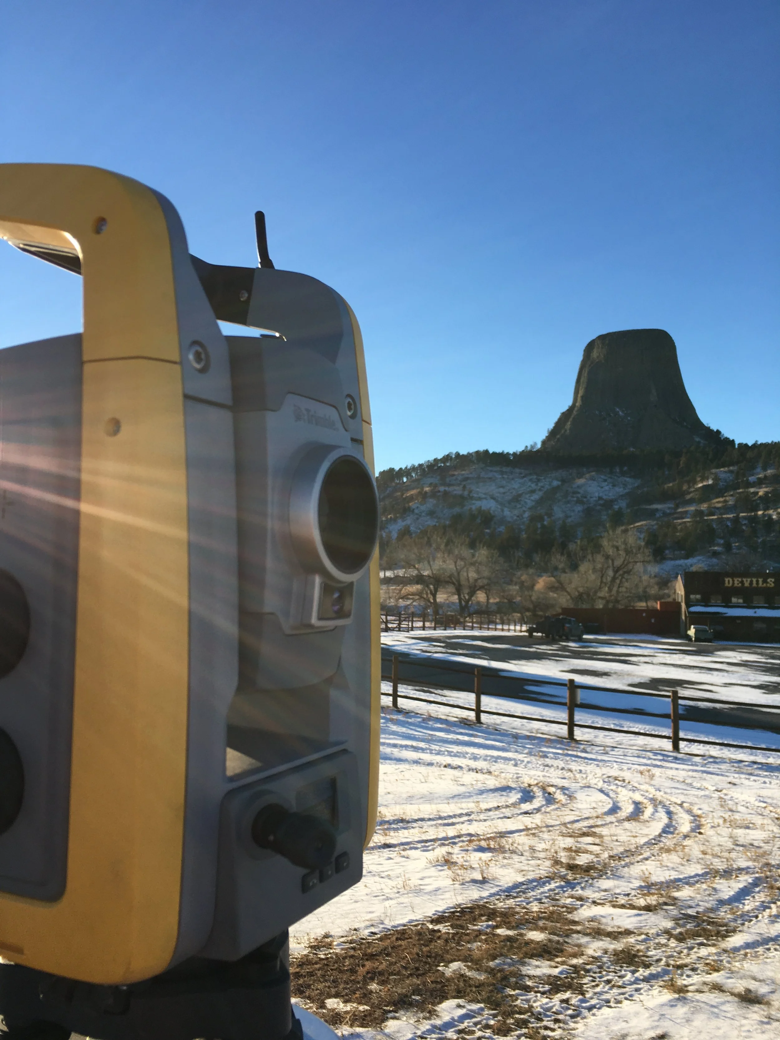 A yellow and gray distant viewing telescope in the foreground, with a mountain in the background, and a snow-covered parking lot with a fence and some vehicles.