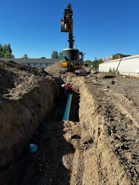 Construction worker installing a blue pipe in a trench with a small excavator nearby under a clear blue sky.