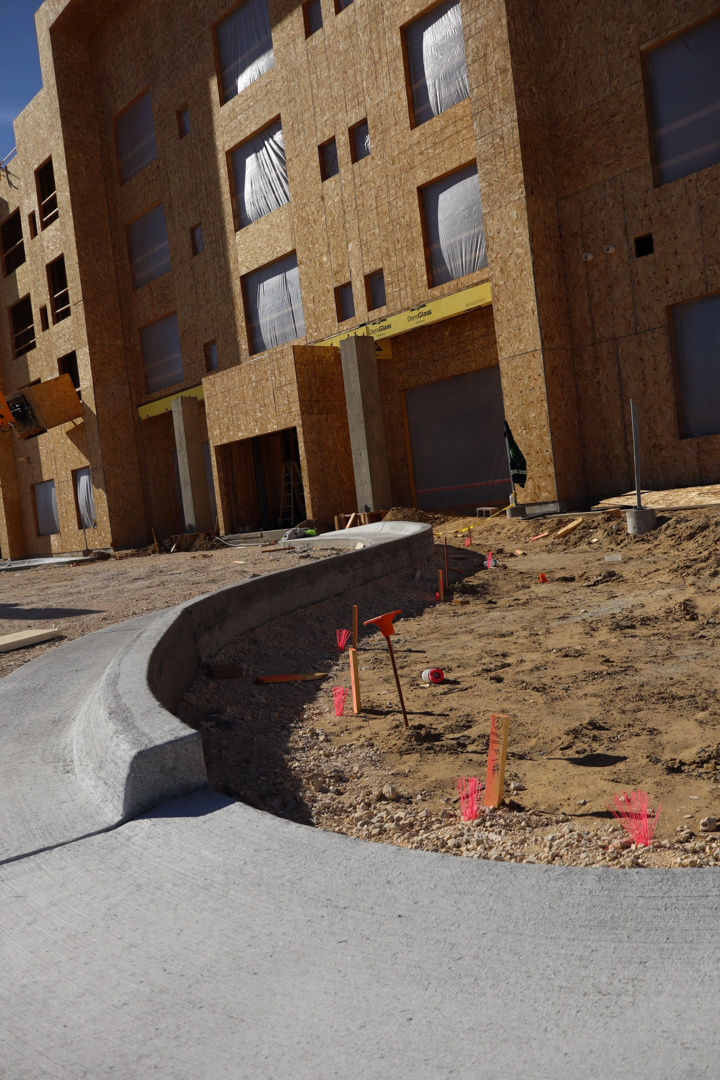 Construction site with a curved concrete sidewalk, wooden building frames with insulation, and various construction tools and markers in the dirt