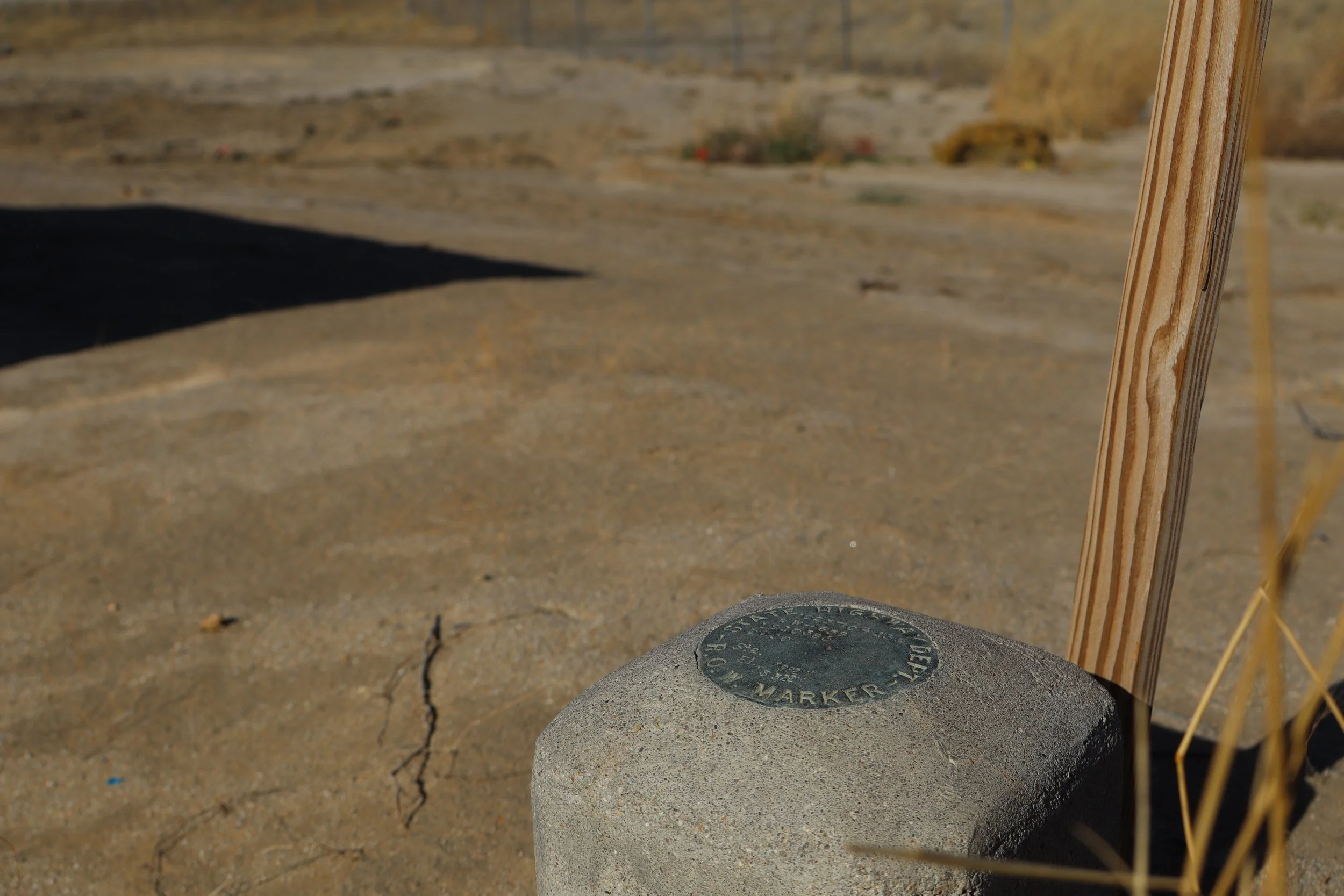 Close-up of a concrete marker with a circular metal plaque on top, placed on sandy ground with cracks. A wooden post is partially visible on the right side, and there are dunes or hills in the background.