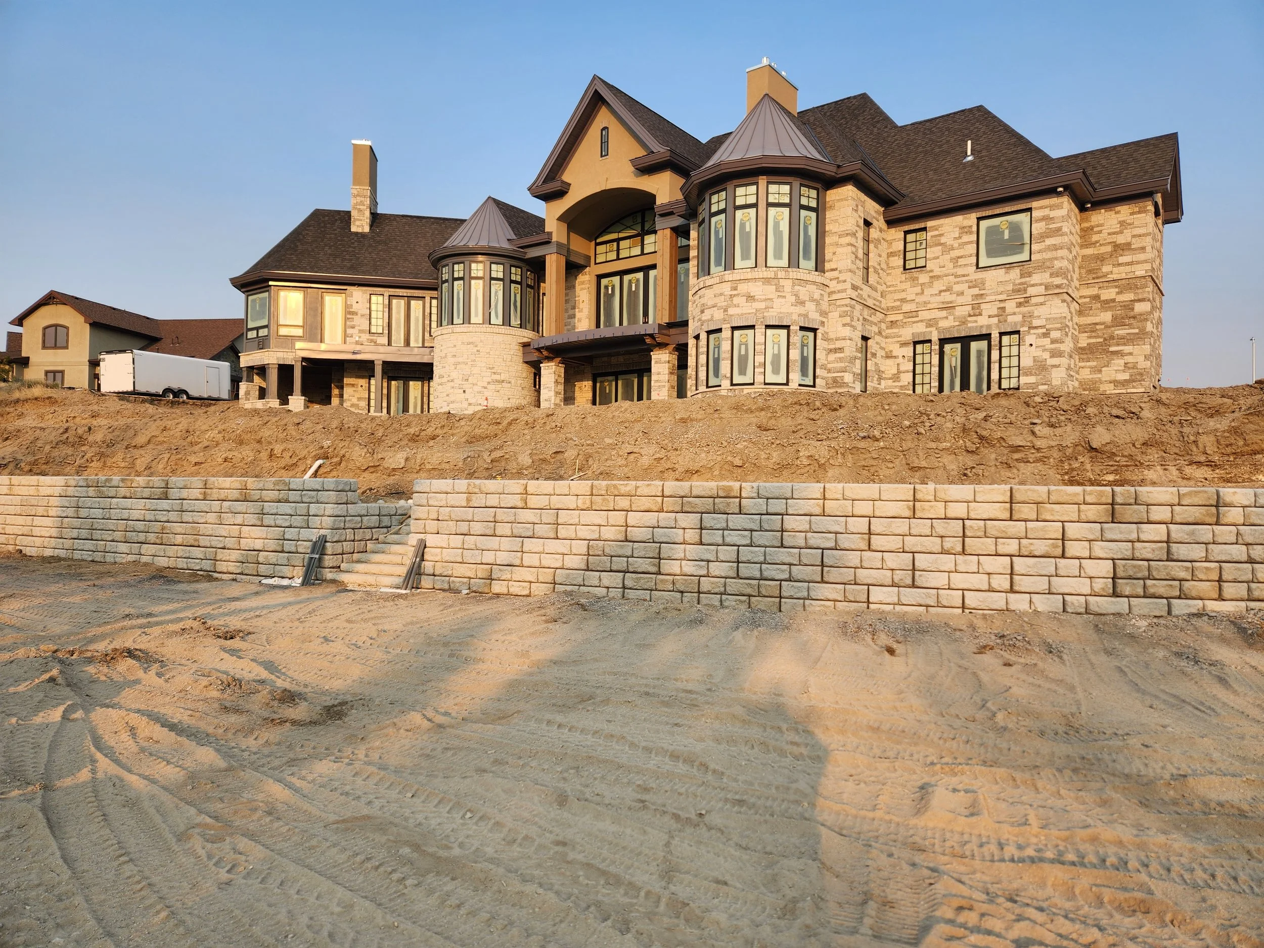 Large two-story brick house under construction with a dirt foreground and a retaining wall, set against a clear sky.