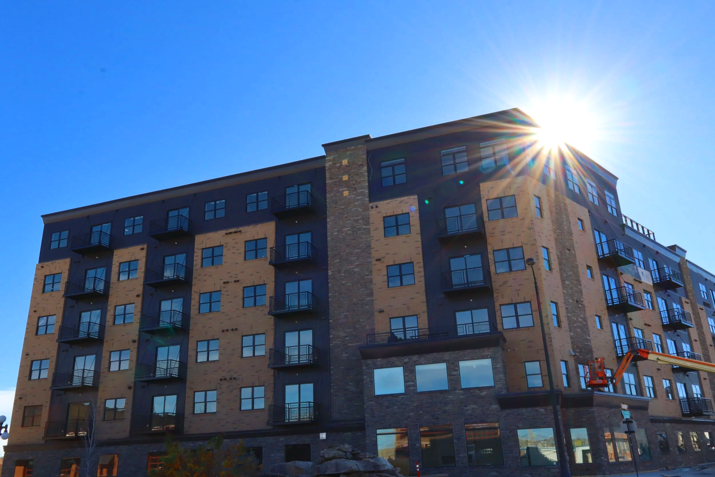 A multi-story residential building with a combination of brick and dark-colored siding, featuring multiple balconies and large windows, with the sun shining brightly behind the building.