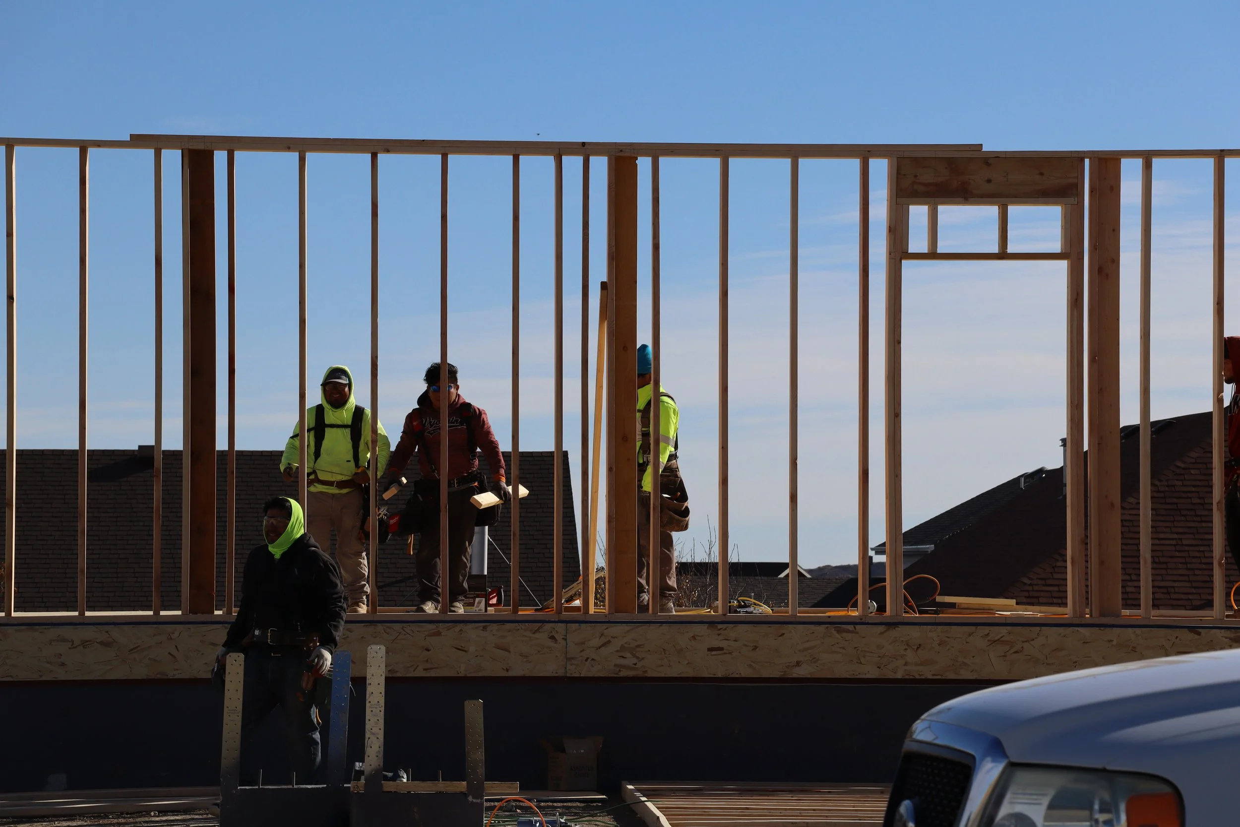 Construction workers working on the wooden frame of a building, with rooftops and a blue sky in the background.