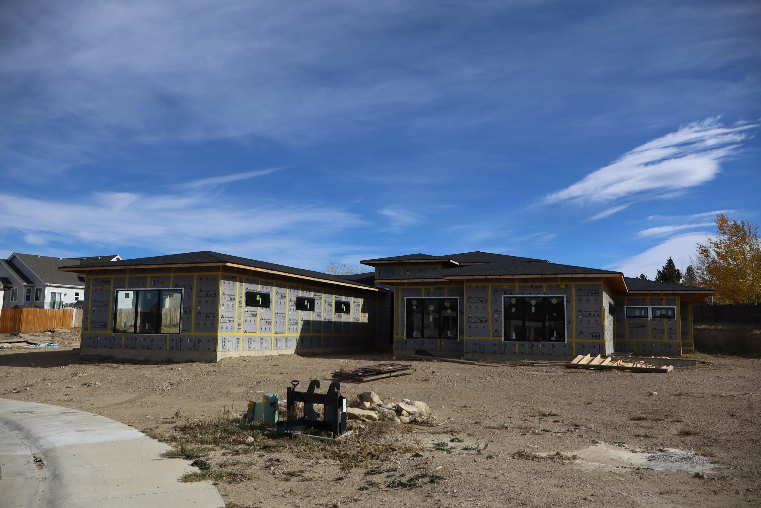 A modern house under construction with black roofing, surrounded by a dirt lot, with neighboring houses in the background and a partly cloudy blue sky overhead.