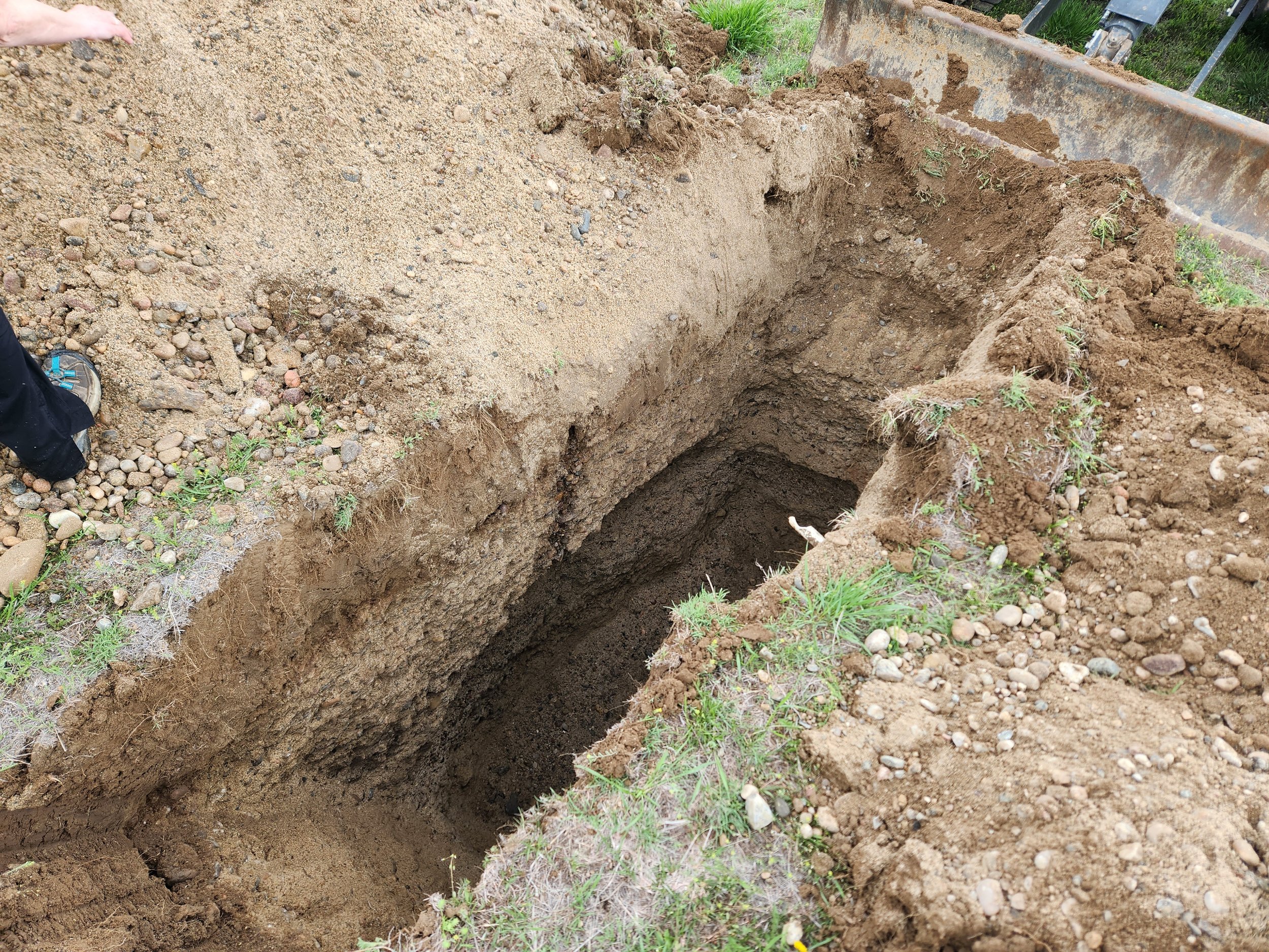 A large, rectangular hole dug into the ground with visible layers of soil. Part of a person’s foot is seen on the left side, wearing a shoe. Green grass and small plants are around the edges of the hole.