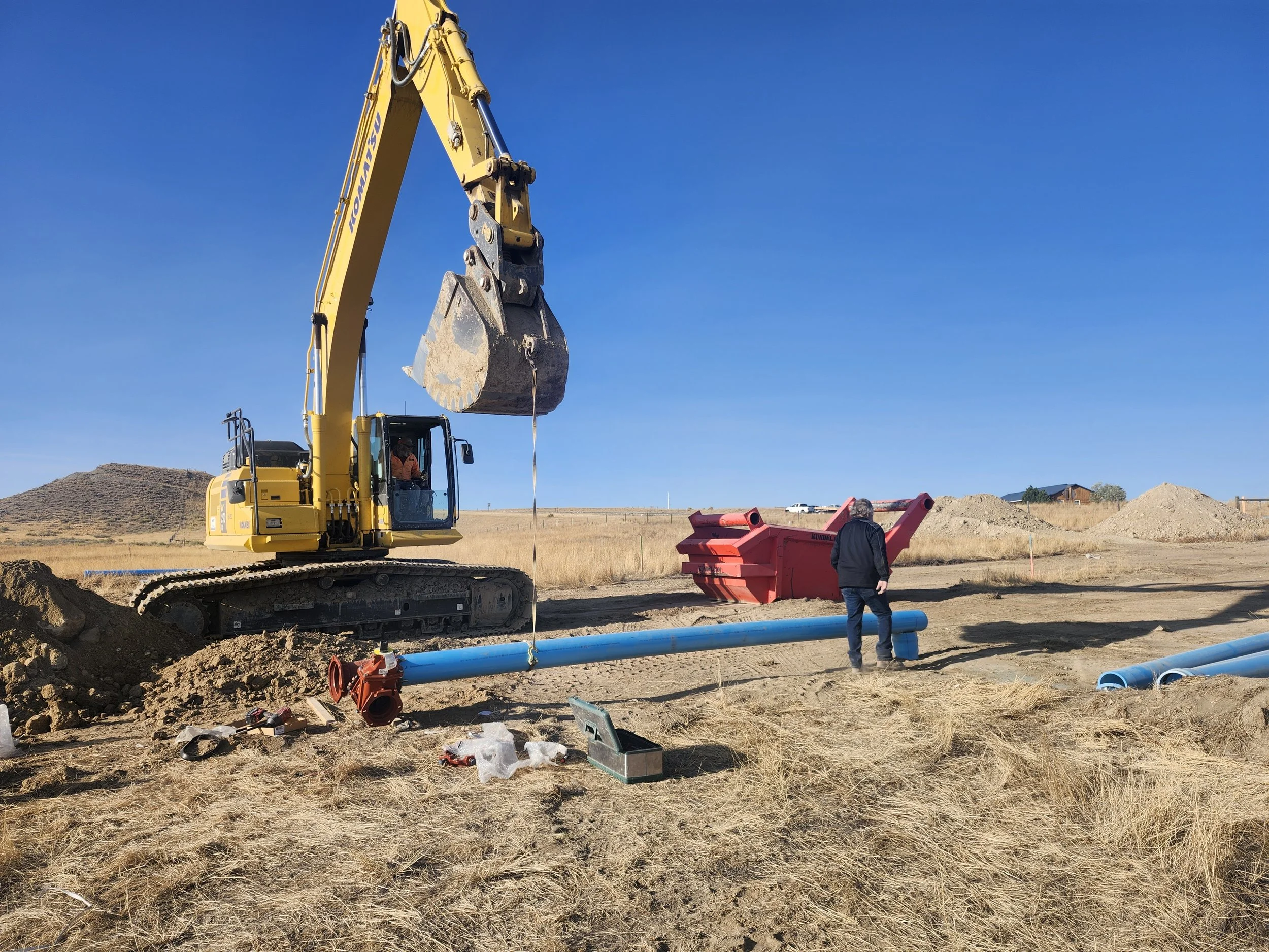 A construction site on a clear day with a yellow excavator digging into the ground near blue pipes. A person in dark clothing stands nearby observing. Mounds of dirt and construction equipment are visible in the background.