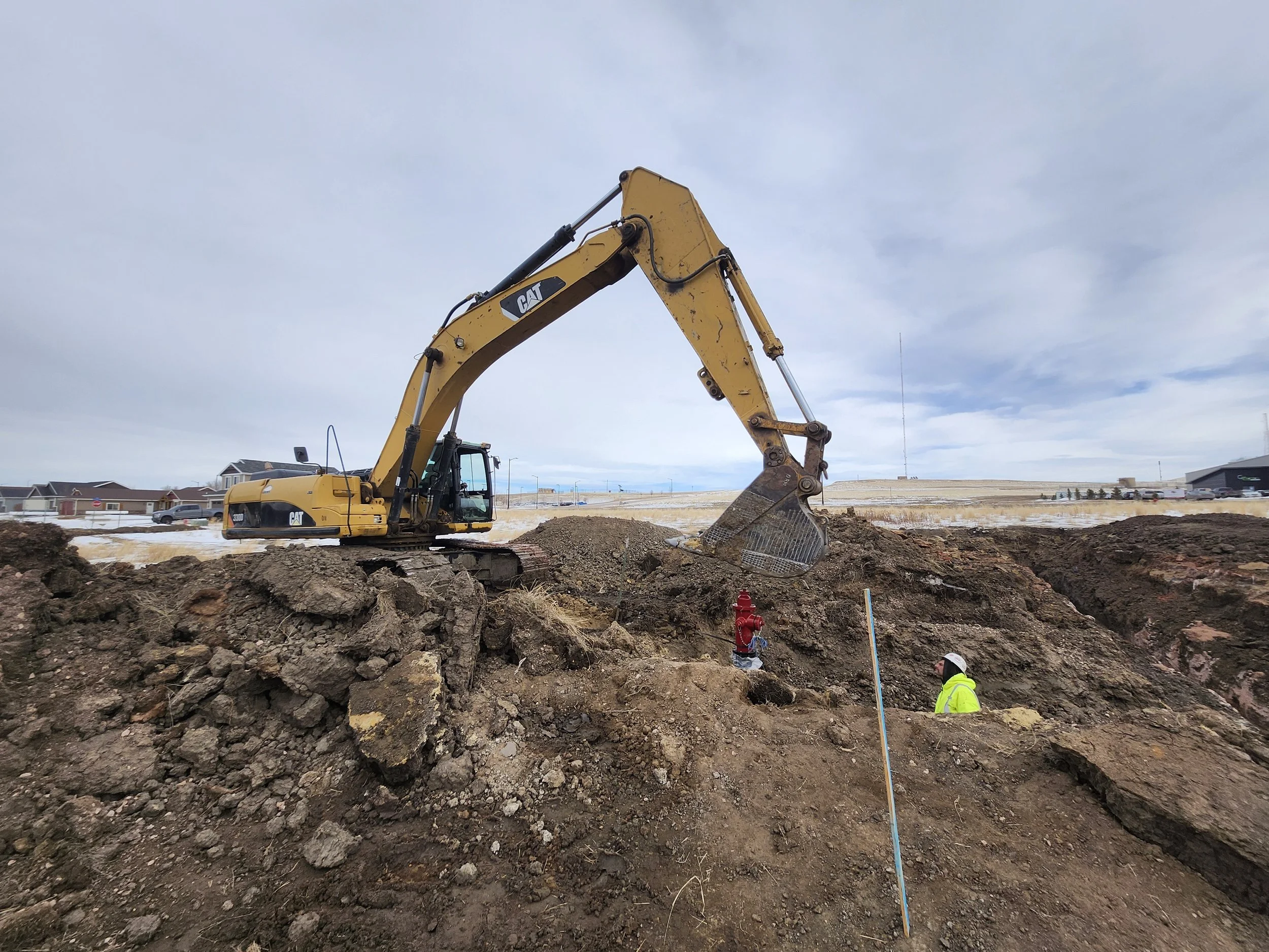 A construction site with a large yellow Caterpillar excavator working on the dirt, with workers in safety gear nearby, near residential homes under a cloudy sky.