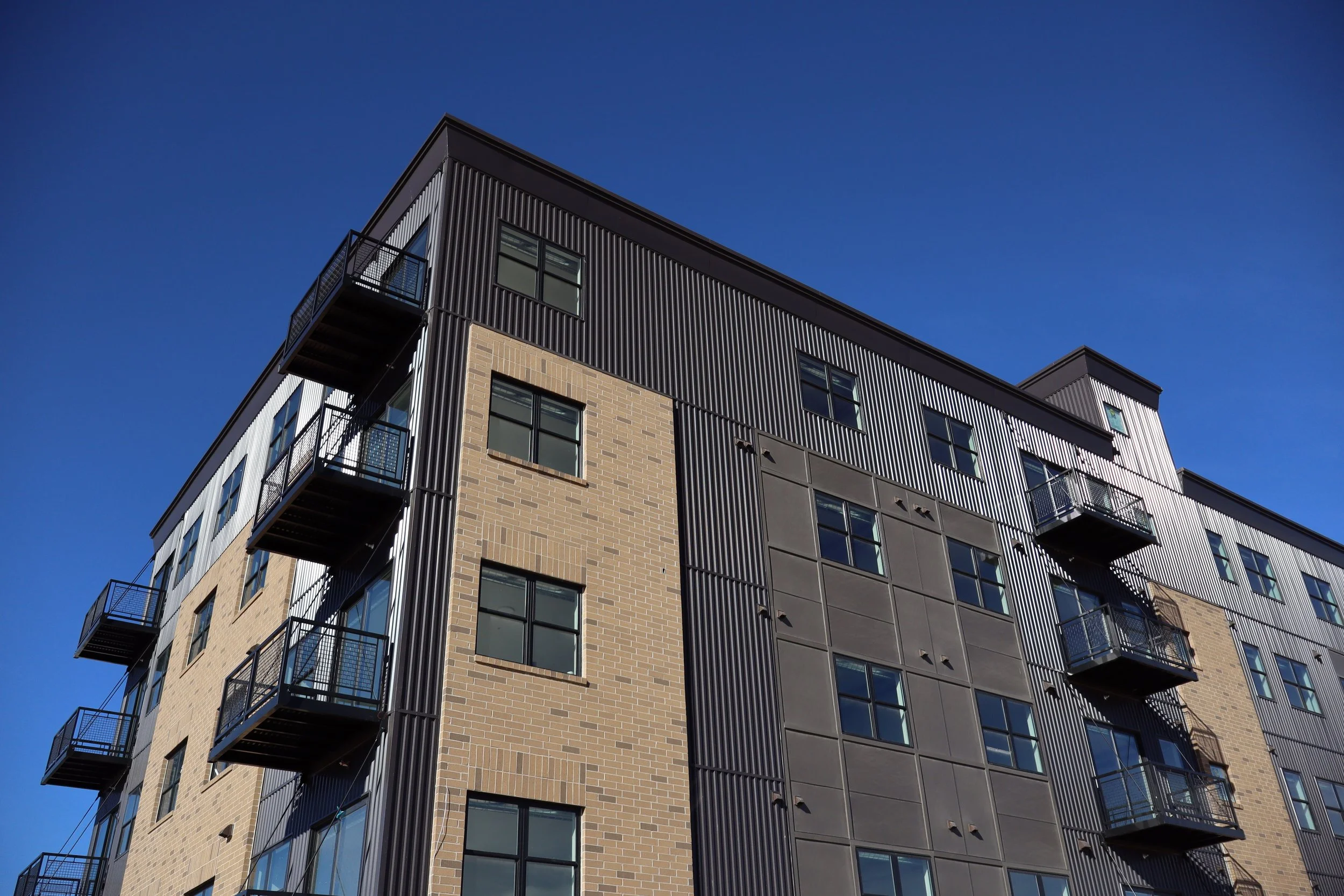 A modern multi-story apartment building with brick and metal panel exterior, featuring glass windows and multiple small balconies against a clear blue sky.