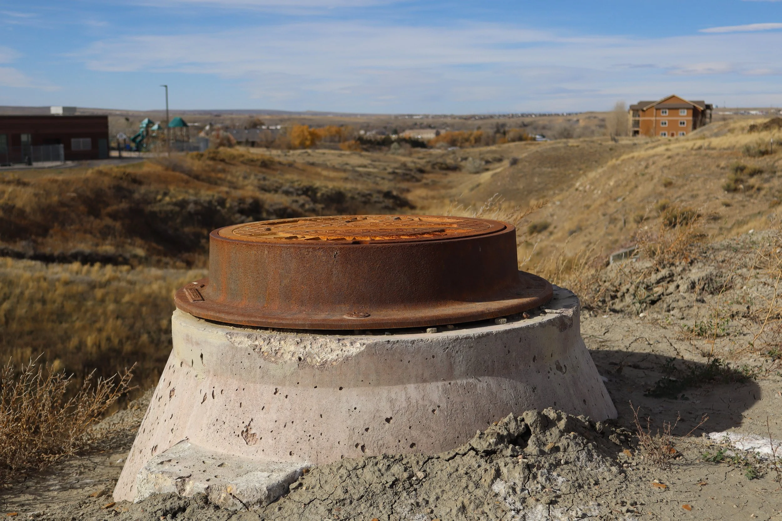 A rusty manhole cover set on a concrete foundation in a dry, hilly landscape under a partly cloudy sky.