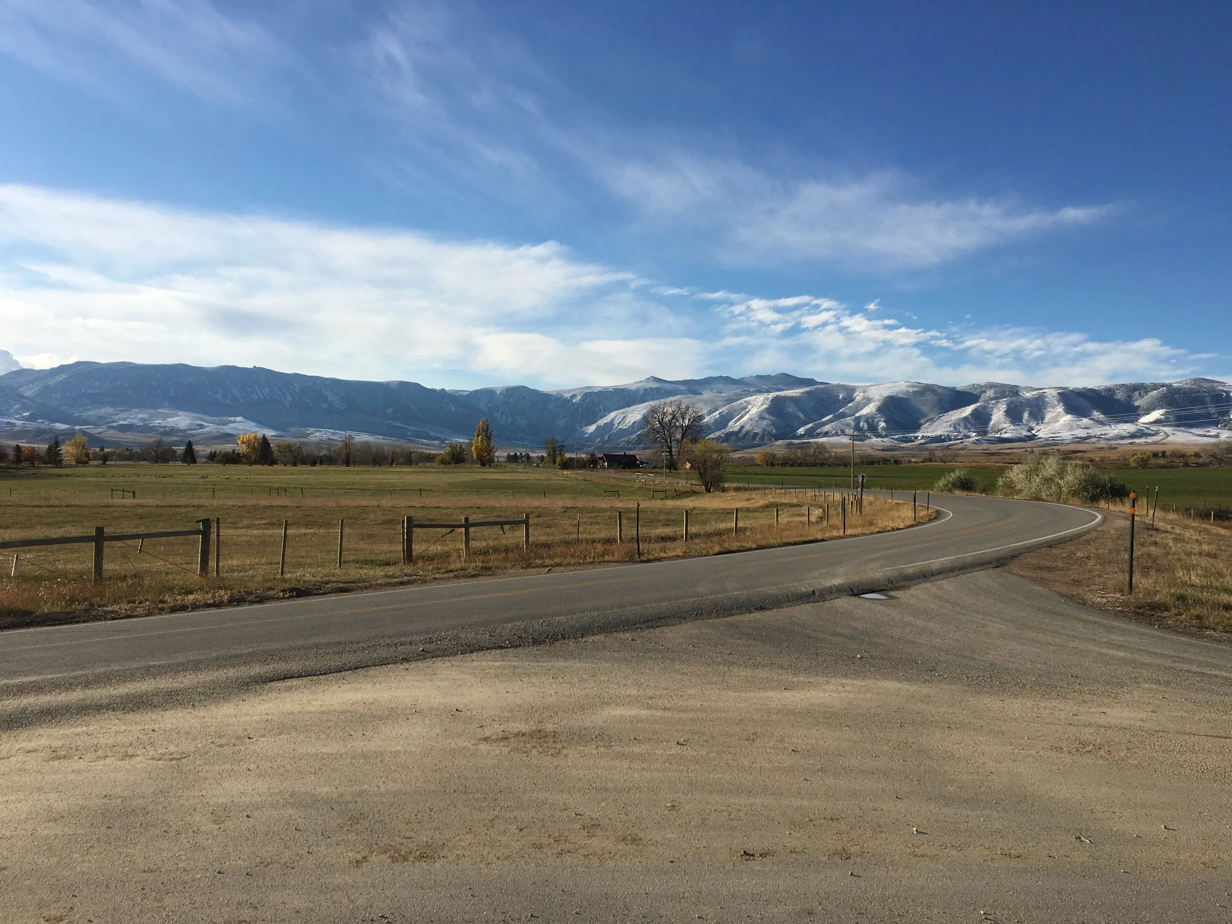 A rural landscape with a curved paved road, grassy fields, and distant snow-capped mountains under a blue sky with scattered clouds.