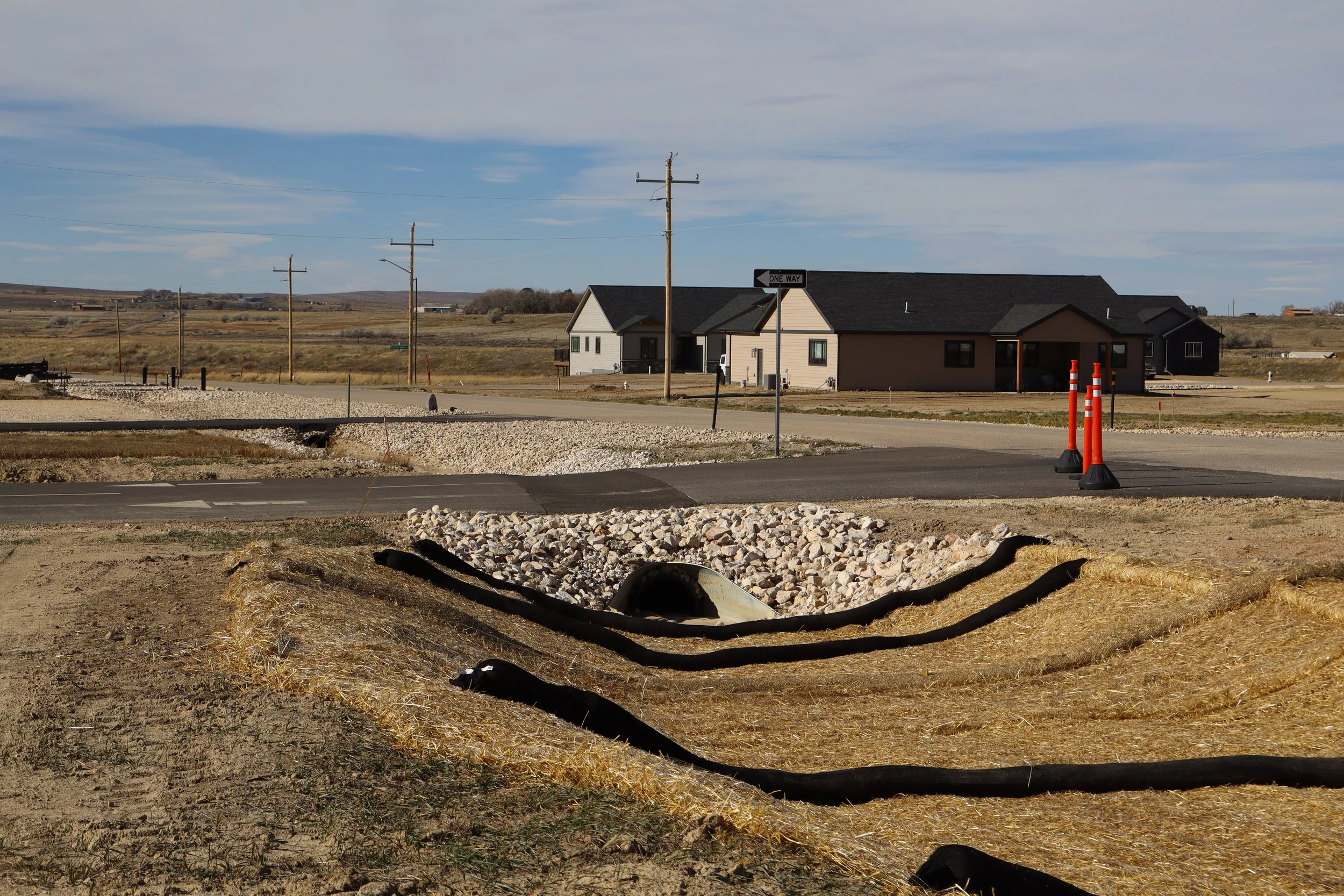 Construction site with new roadwork, orange traffic cones, gravel, and a residential house in the background under a partly cloudy sky.