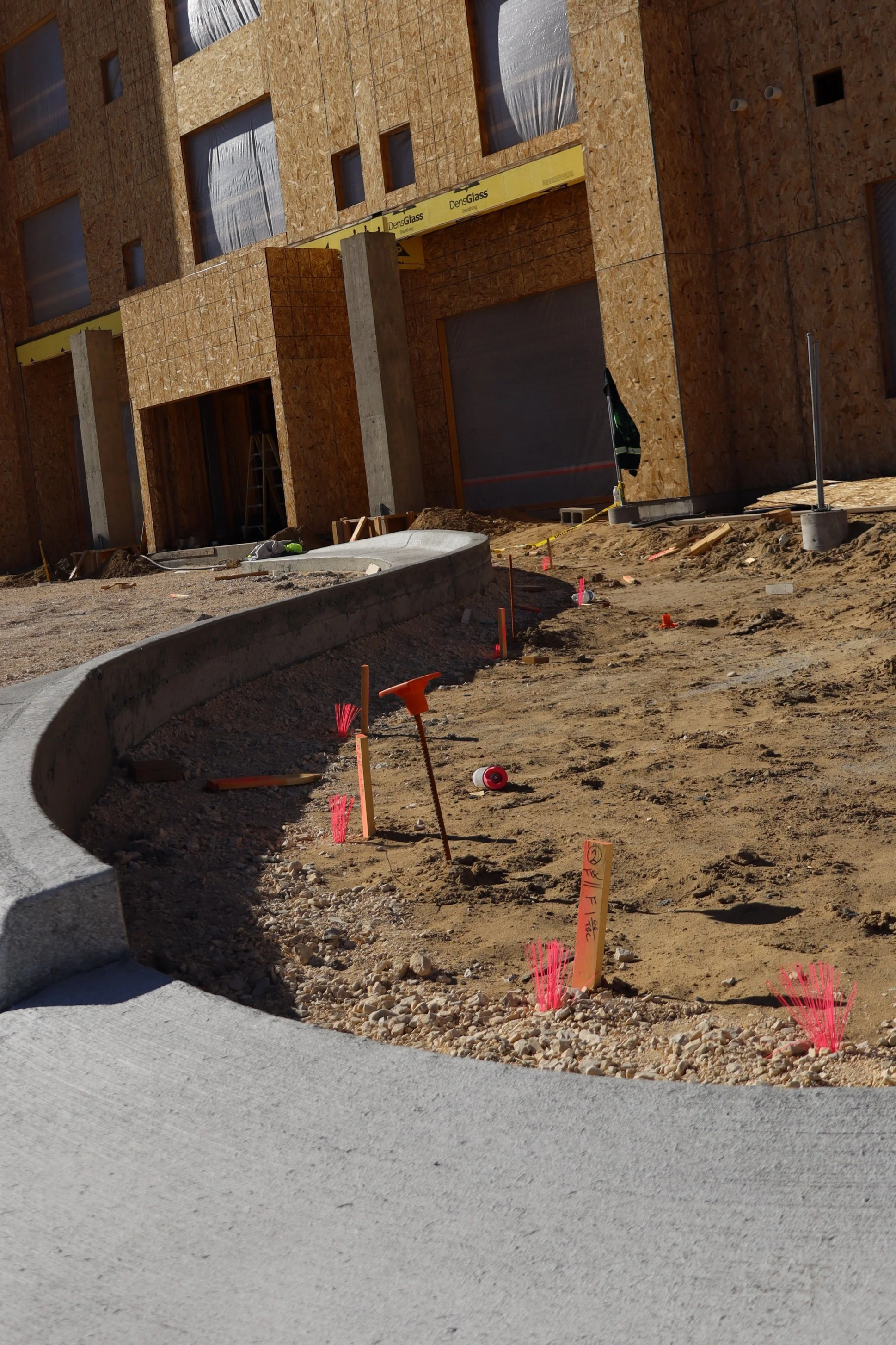 Construction site of a building with exposed plywood walls, a curved concrete pathway, and construction markers and tools on the dirt ground.