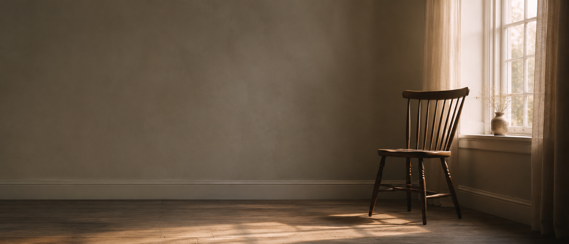 Empty wooden chair placed near a window with curtains and a small vase with dried flowers, sunlight streaming in, in a minimalist room.