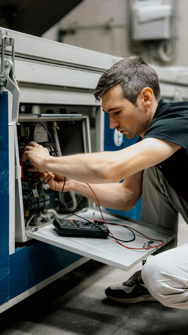 A young man working on an electrical panel with a multimeter connected to it, in an industrial or technical setting.