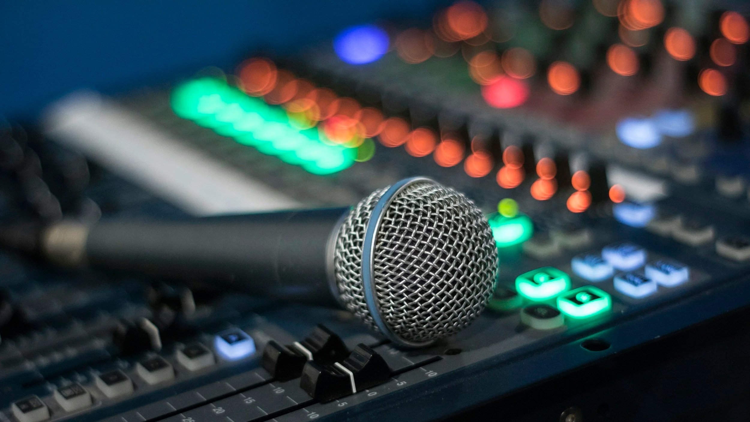 A close-up of a silver microphone placed on a sound mixer with colorful illuminated controls and sliders, used for audio recording or podcasting.