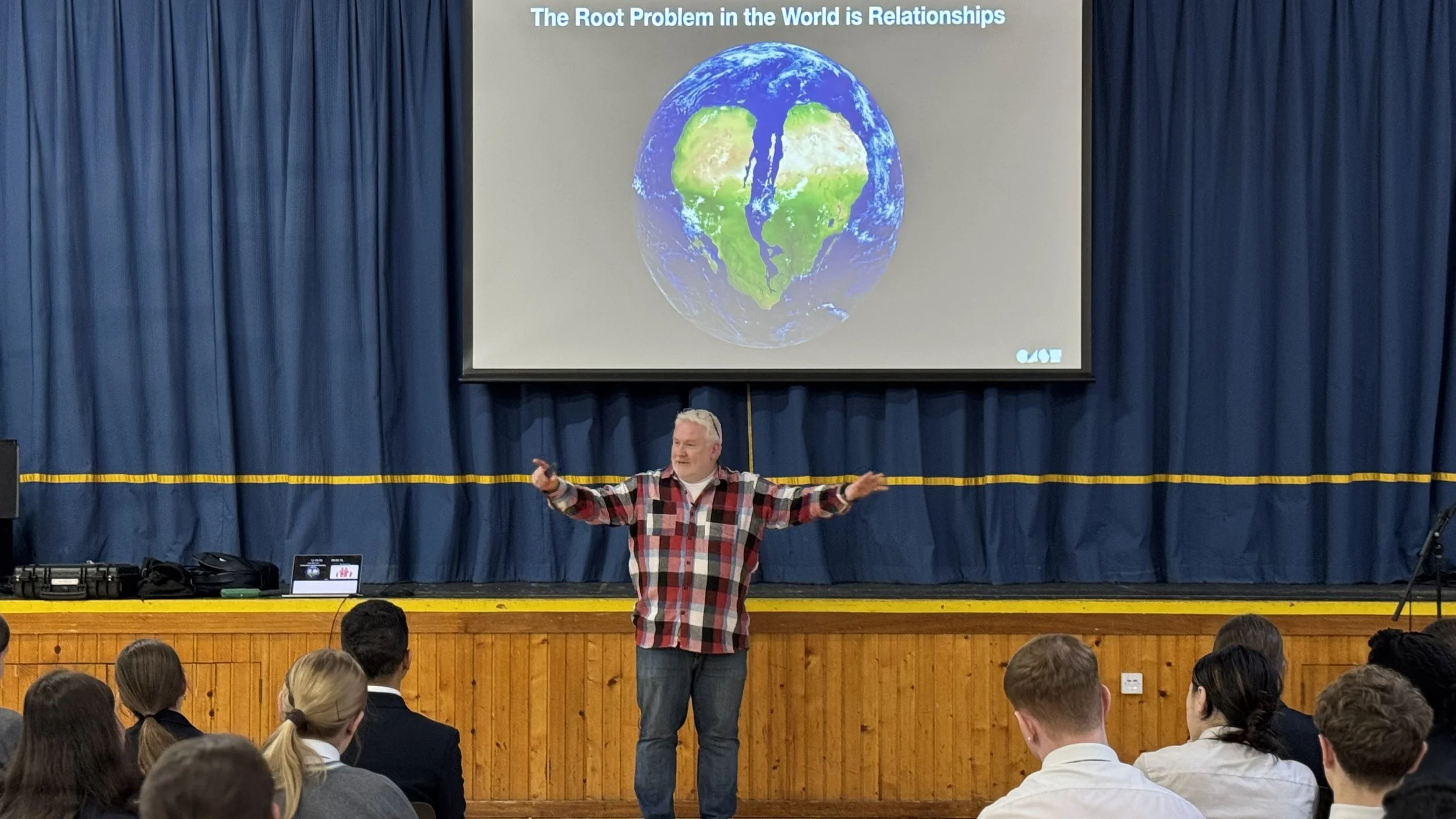 A man in a red and black plaid shirt is giving a presentation to an audience in a large auditorium. He is standing in front of a blue curtain with a projection screen behind him displaying an image of the Earth with the continents arranged to form a heart shape. The slide has the text "The Root Problem in the World is Relationships" at the top.