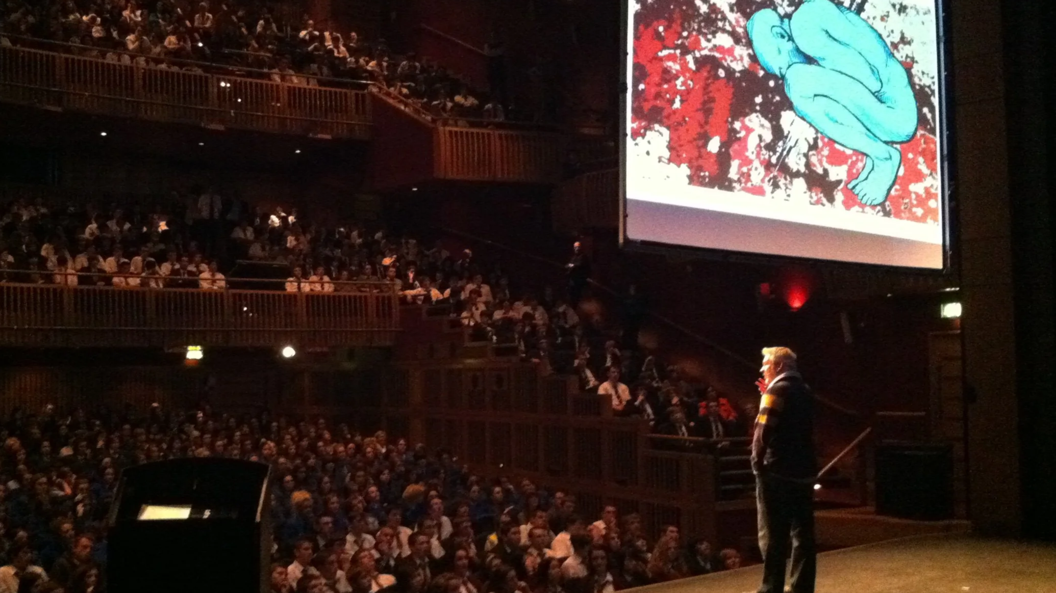 Large auditorium filled with many seated audience members, stage with a speaker at podium, large screen showing artwork of a blue figure in a crouched position on a splattered red and black background.