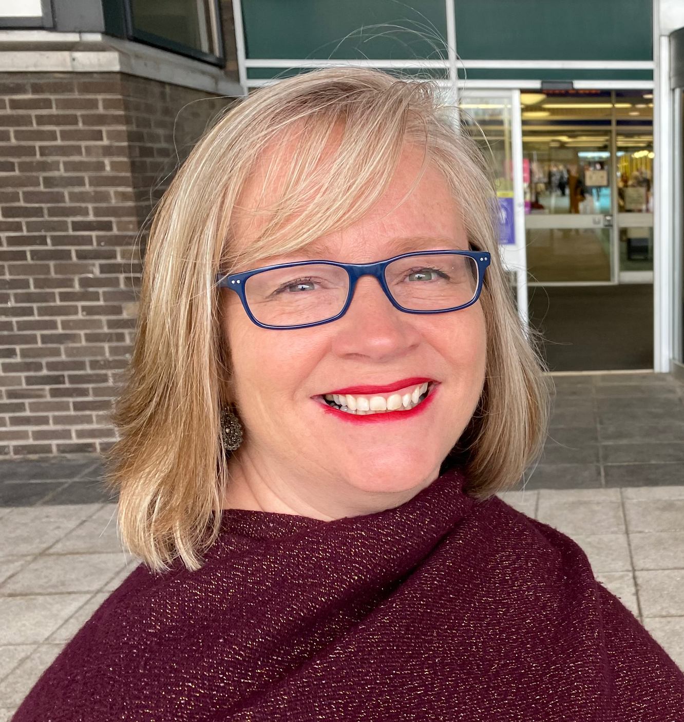 A woman smiling outdoors in front of a building with glass doors and brick walls, wearing glasses, a maroon shawl, and red lipstick.