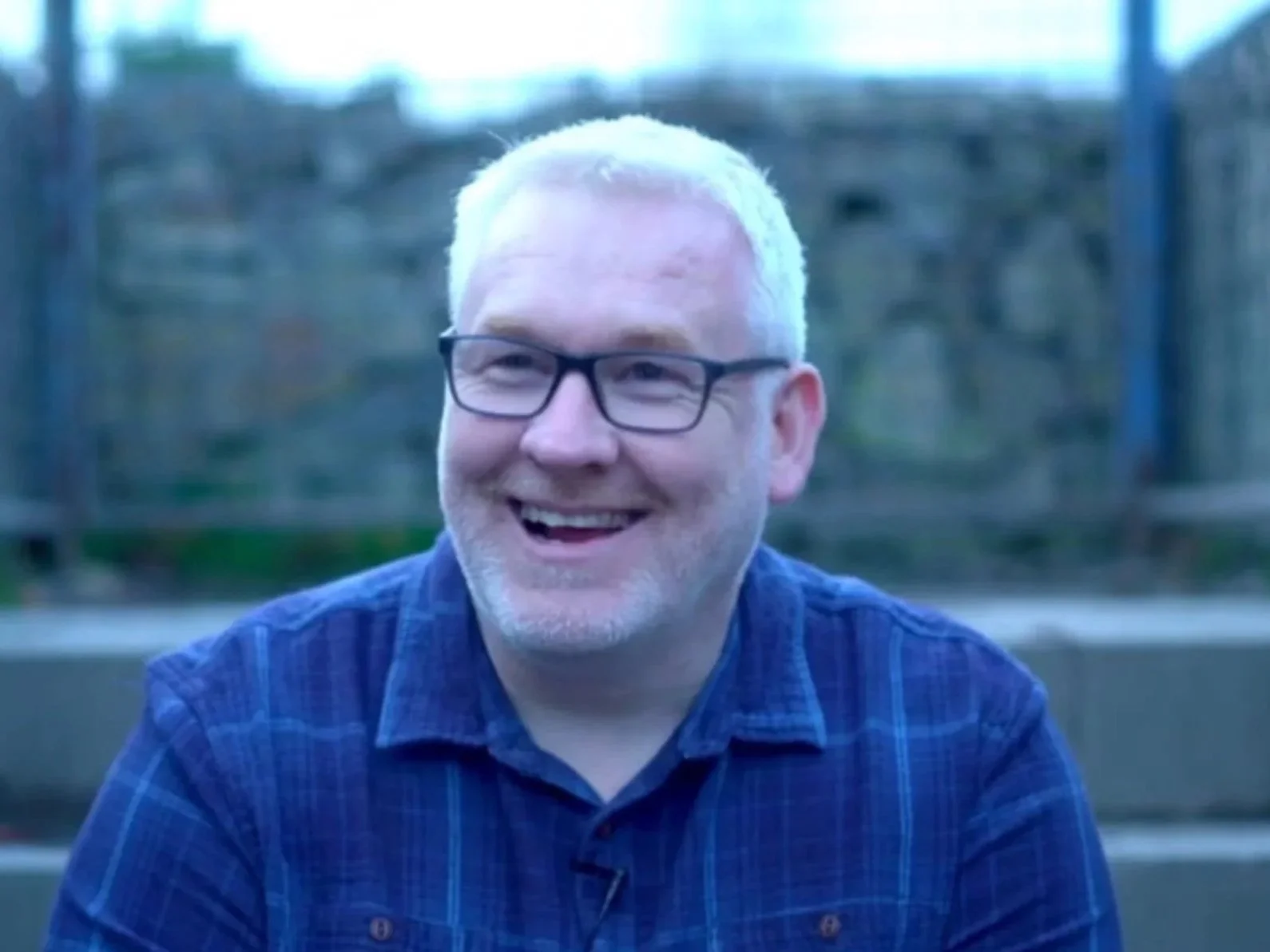 A smiling man with glasses and white hair sits outdoors, wearing a blue checkered shirt.