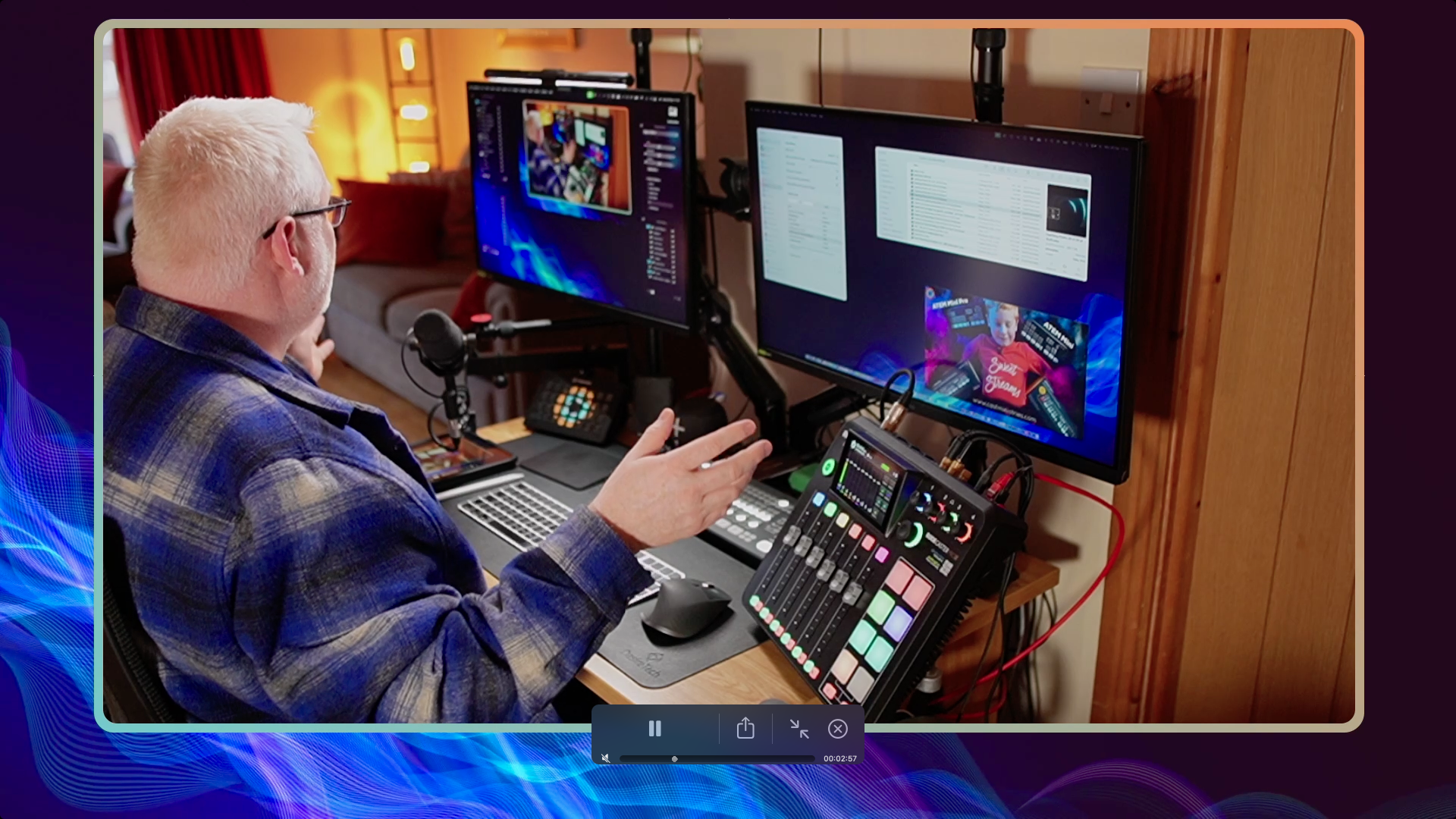 Man with white hair and glasses working at a home studio setup with multiple monitors, audio equipment, and a microphone.