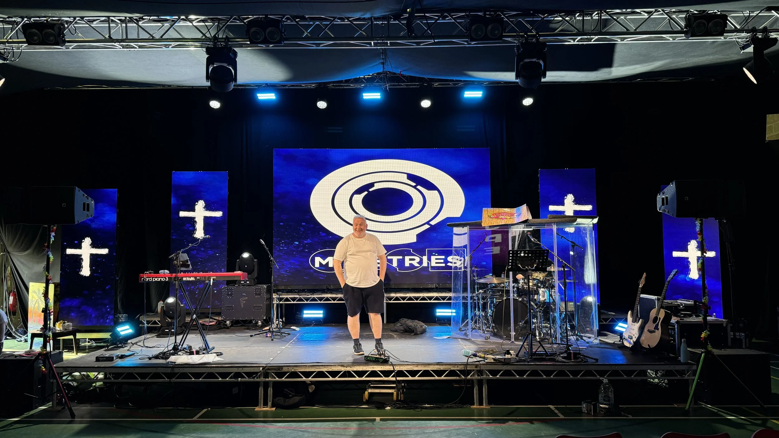 A man standing on a stage in front of a large screen displaying the logo 'MOTRES'. The stage has musical instruments, microphones, and equipment, with crosses and blue lighting in the background, indicating a religious or worship event.
