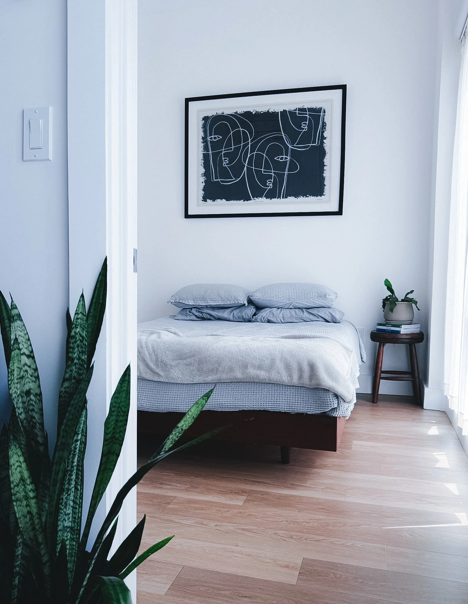 A bedroom with a bed against a white wall, framed abstract line art above, a small round side table with books and a potted plant to the right, and a tall indoor plant with long green leaves on the left.