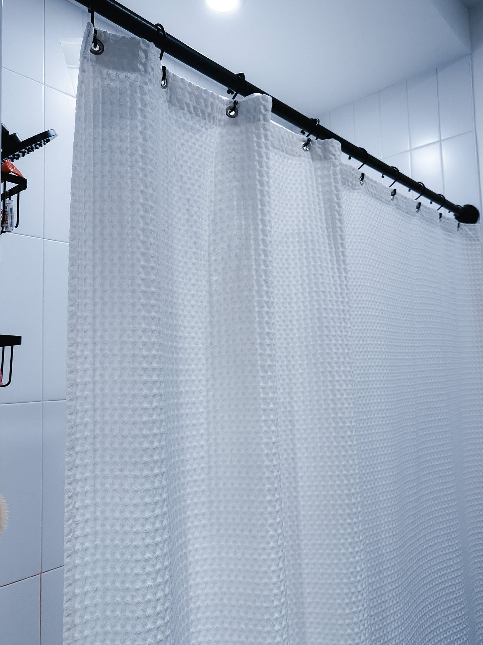 White textured shower curtain hanging from a black metal rod in a bathroom with white tiled walls.