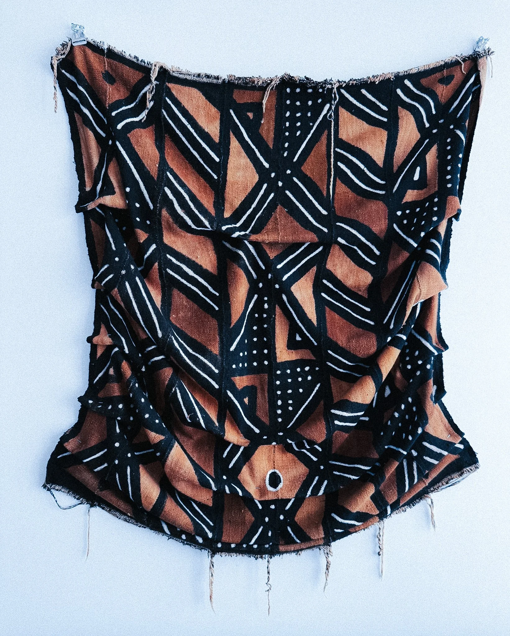 Colorful tribal-patterned fabric wall hanging with black, orange, and white geometric designs, frayed edges, and hanging strings.