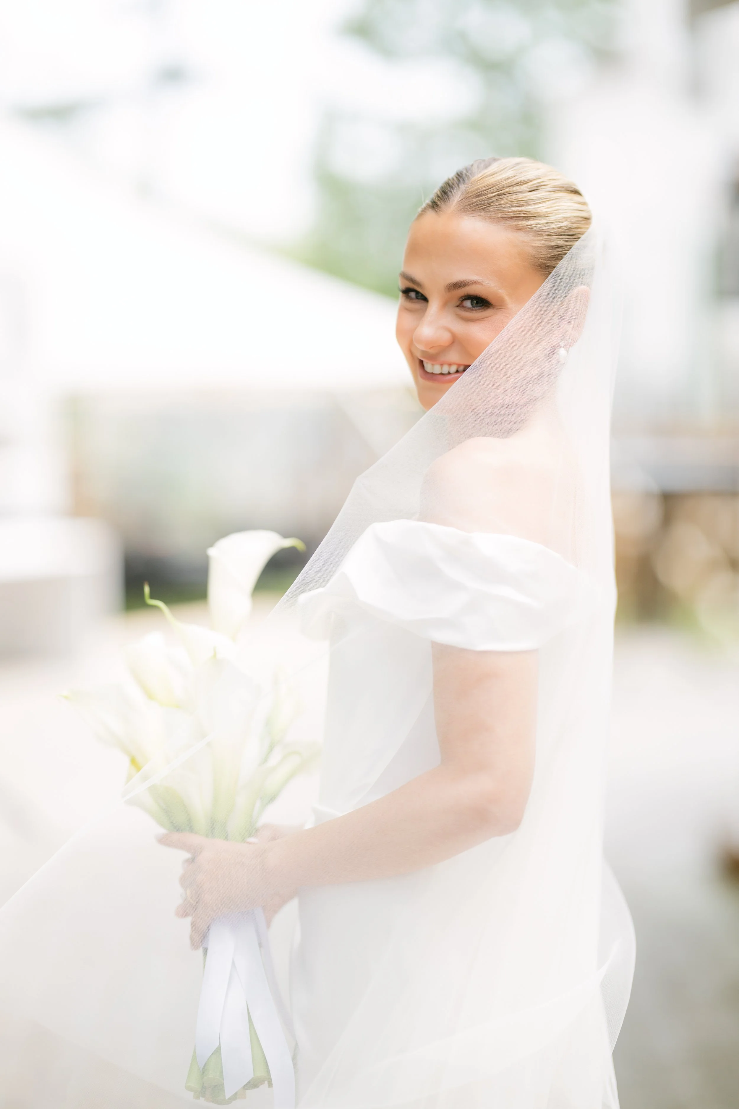 A smiling bride with a veil and pearl earrings holds a bouquet of white lilies.