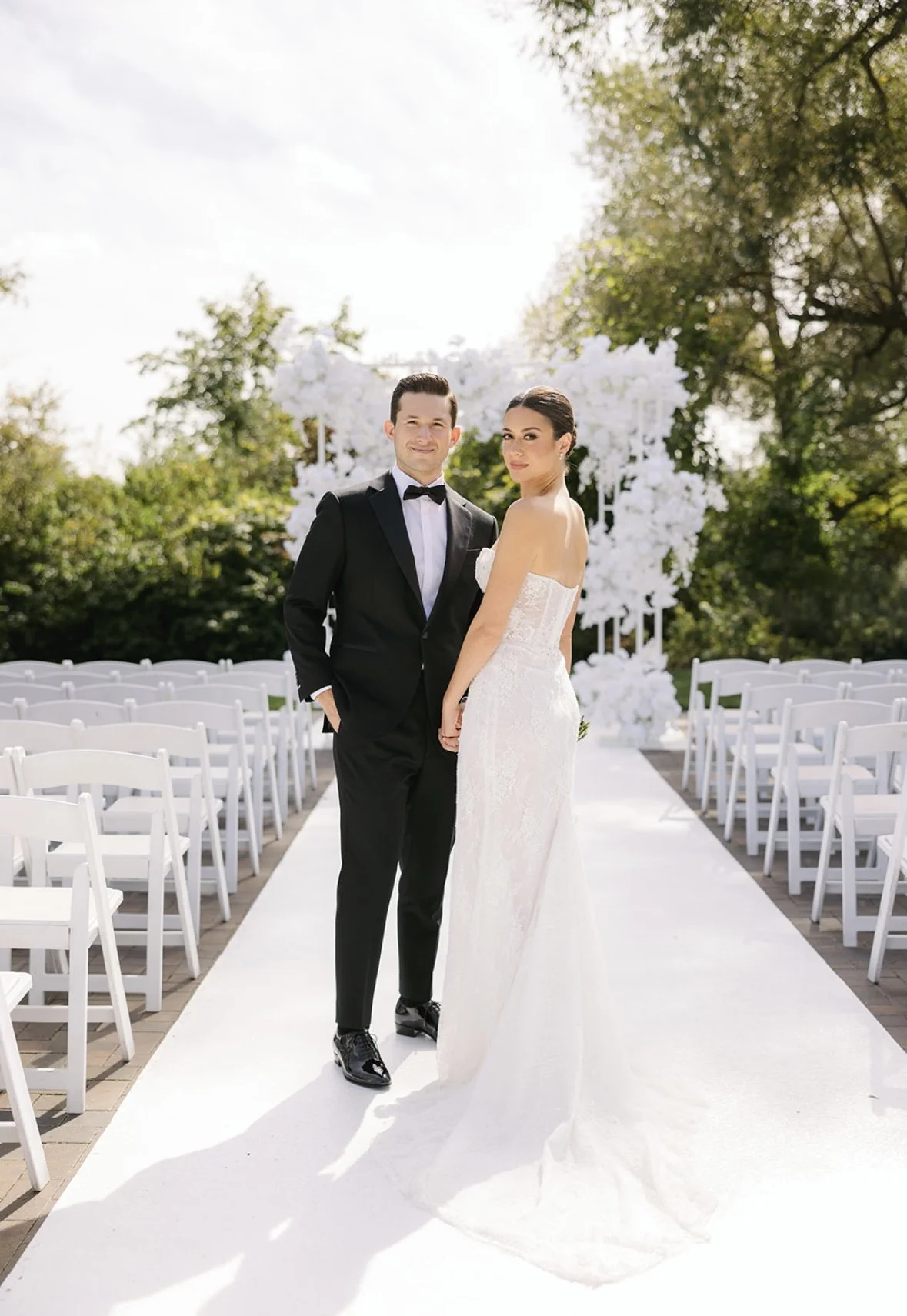 A bride and groom standing outdoors on a wedding aisle, with white chairs and floral arch in the background.